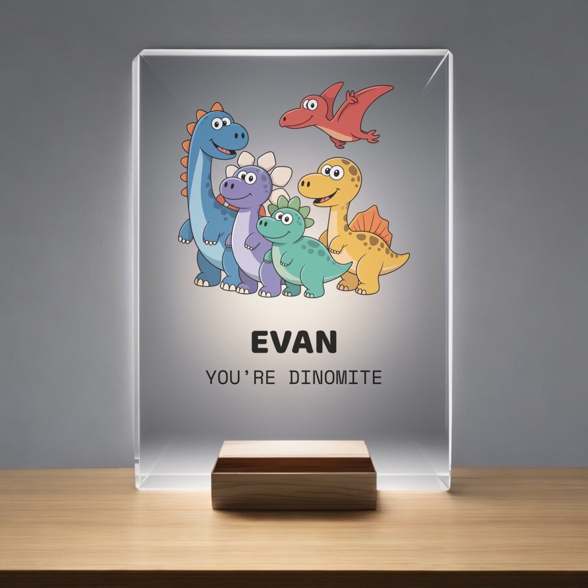 The Mirabilia Boutique Personalized Dinosaur Night Light for Kids features a glass plaque with cartoon dinosaurs, "Evan," and "You're dinomite," displayed on a wooden stand—ideal for any child's bedroom night light collection.