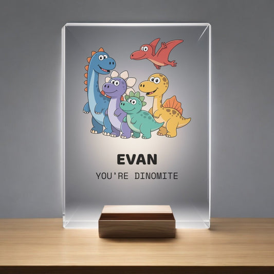 The Mirabilia Boutique Personalized Dinosaur Night Light for Kids features a glass plaque with cartoon dinosaurs, "Evan," and "You're dinomite," displayed on a wooden stand—ideal for any child's bedroom night light collection.