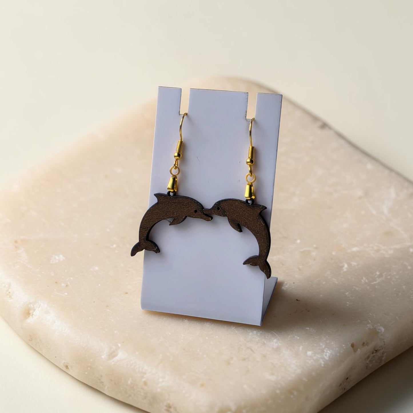 The Dolphin Earrings | Lasercut Walnut Wood by Mirabilia Boutique are displayed on a white stand atop a beige stone, with the earrings facing each other for a symmetrical, ocean-inspired charm.