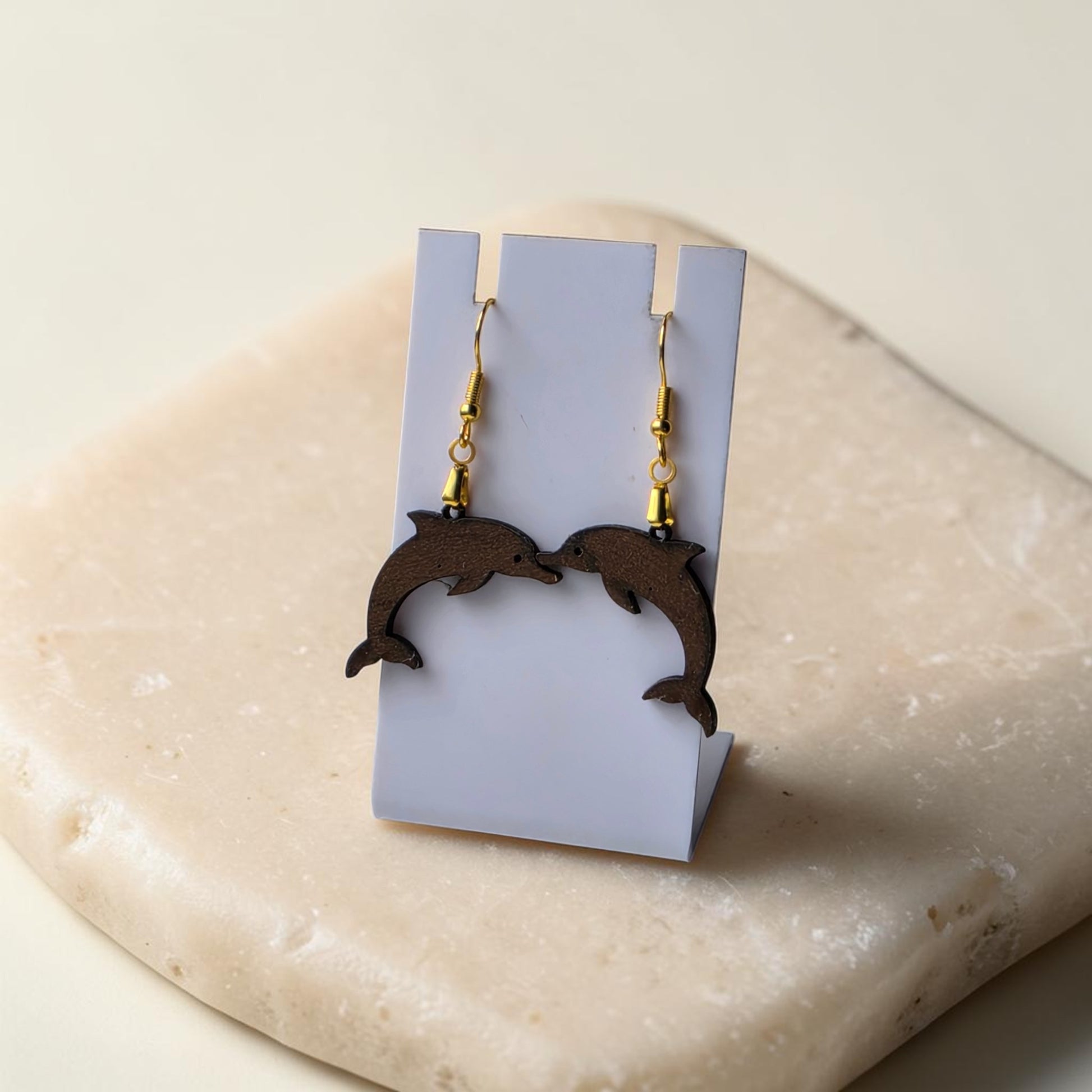 The Dolphin Earrings | Lasercut Walnut Wood by Mirabilia Boutique are displayed on a white stand atop a beige stone, with the earrings facing each other for a symmetrical, ocean-inspired charm.