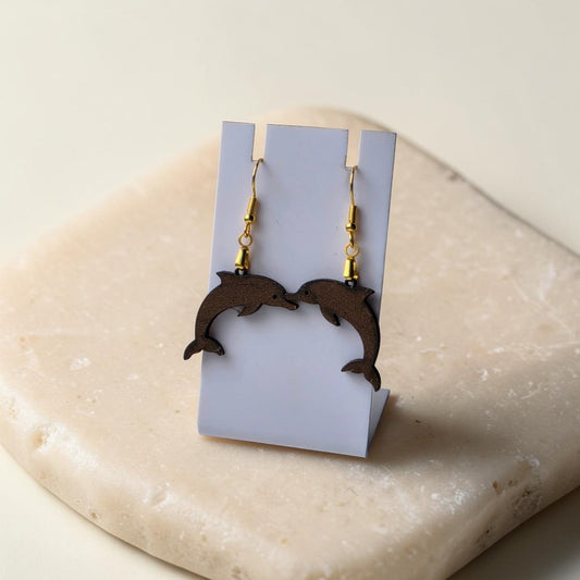 The Dolphin Earrings | Lasercut Walnut Wood by Mirabilia Boutique are displayed on a white stand atop a beige stone, with the earrings facing each other for a symmetrical, ocean-inspired charm.