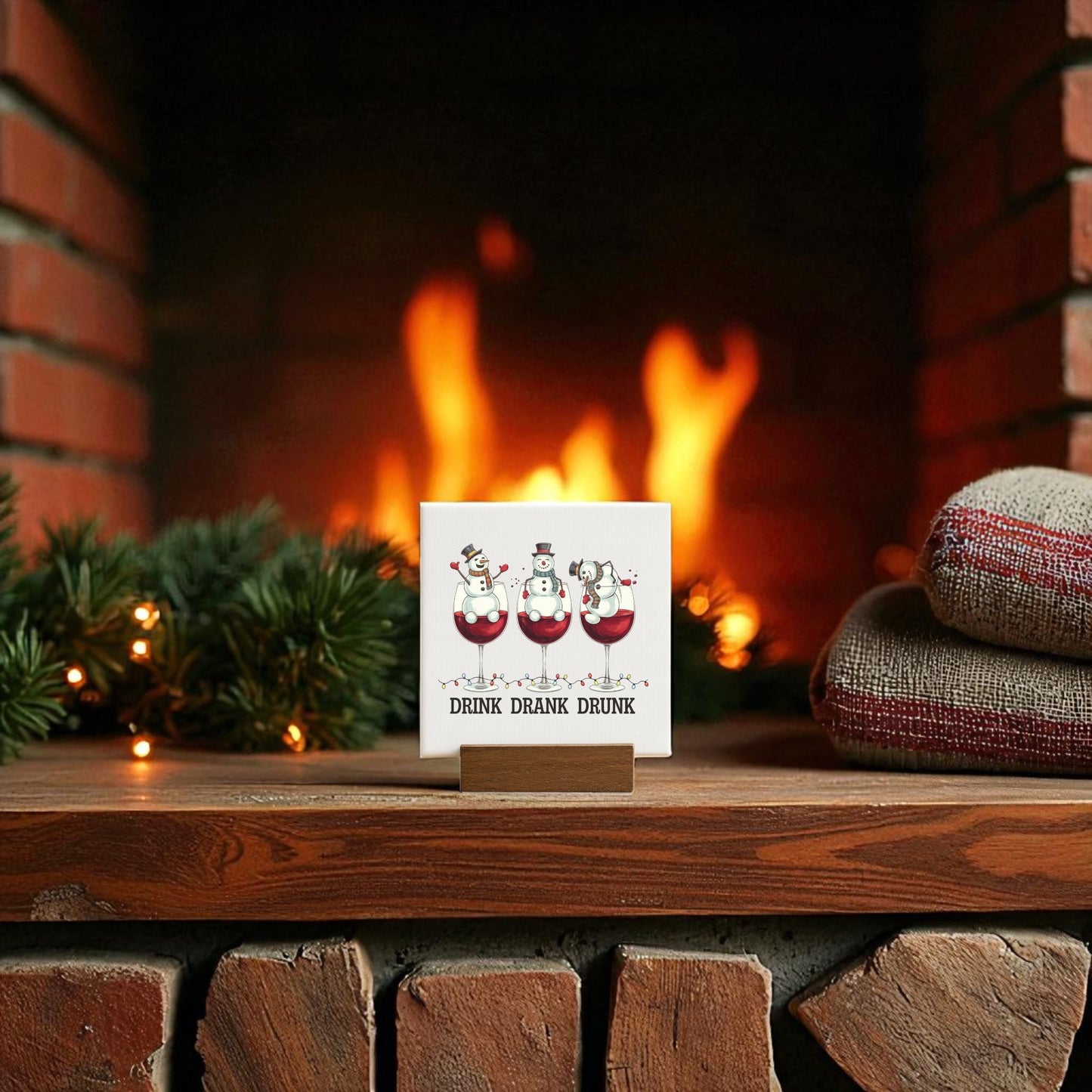 The Drink Drank Drunk Mini Canvas (4x4) by Mirabilia Boutique features cartoon snowmen with wine glasses and festive wording, shown on a wooden mantel with holiday decor; a cozy fireplace and cushions complete the merry scene.