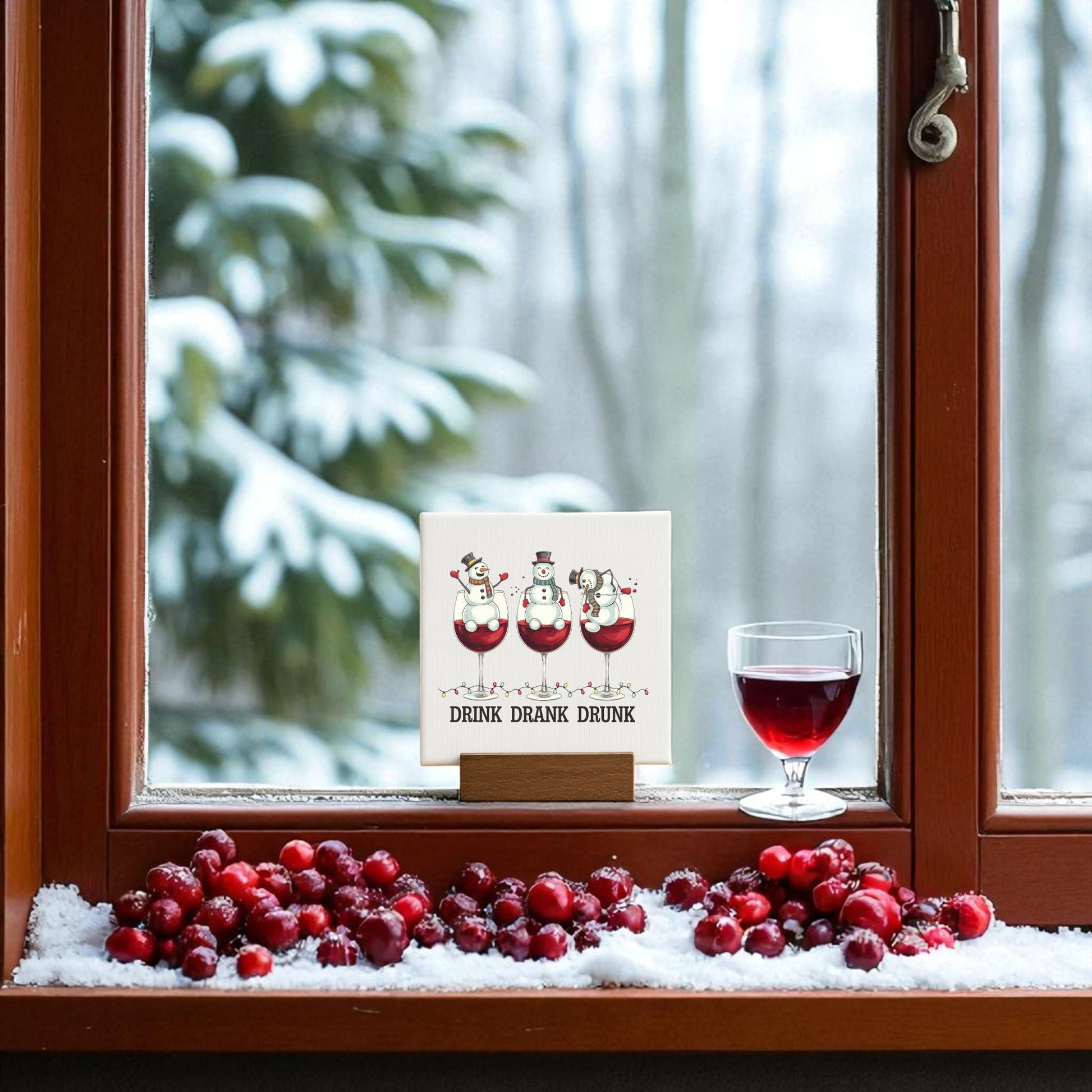 A snowy window ledge displays cranberries, a glass of red wine, and the Mirabilia Boutique "Drink Drank Drunk Mini Canvas" 4x4 funny Christmas sign with three toasting snowmen—festive decor and a perfect gift for any wine lover.