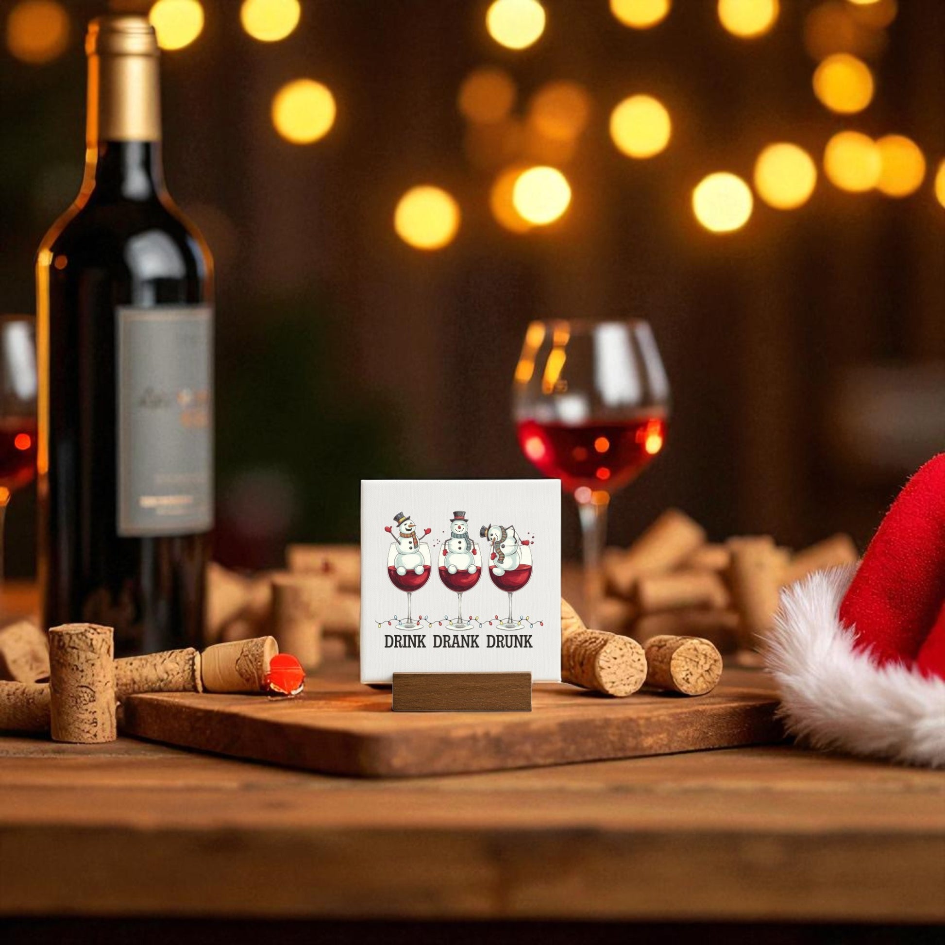 A festive table with wine, two glasses of red, a Santa hat, and Mirabilia Boutique’s "Drink Drank Drunk" 4x4 Mini Canvas—a funny Christmas sign—creates the perfect gift for wine lovers amid warm, glowing holiday lights.
