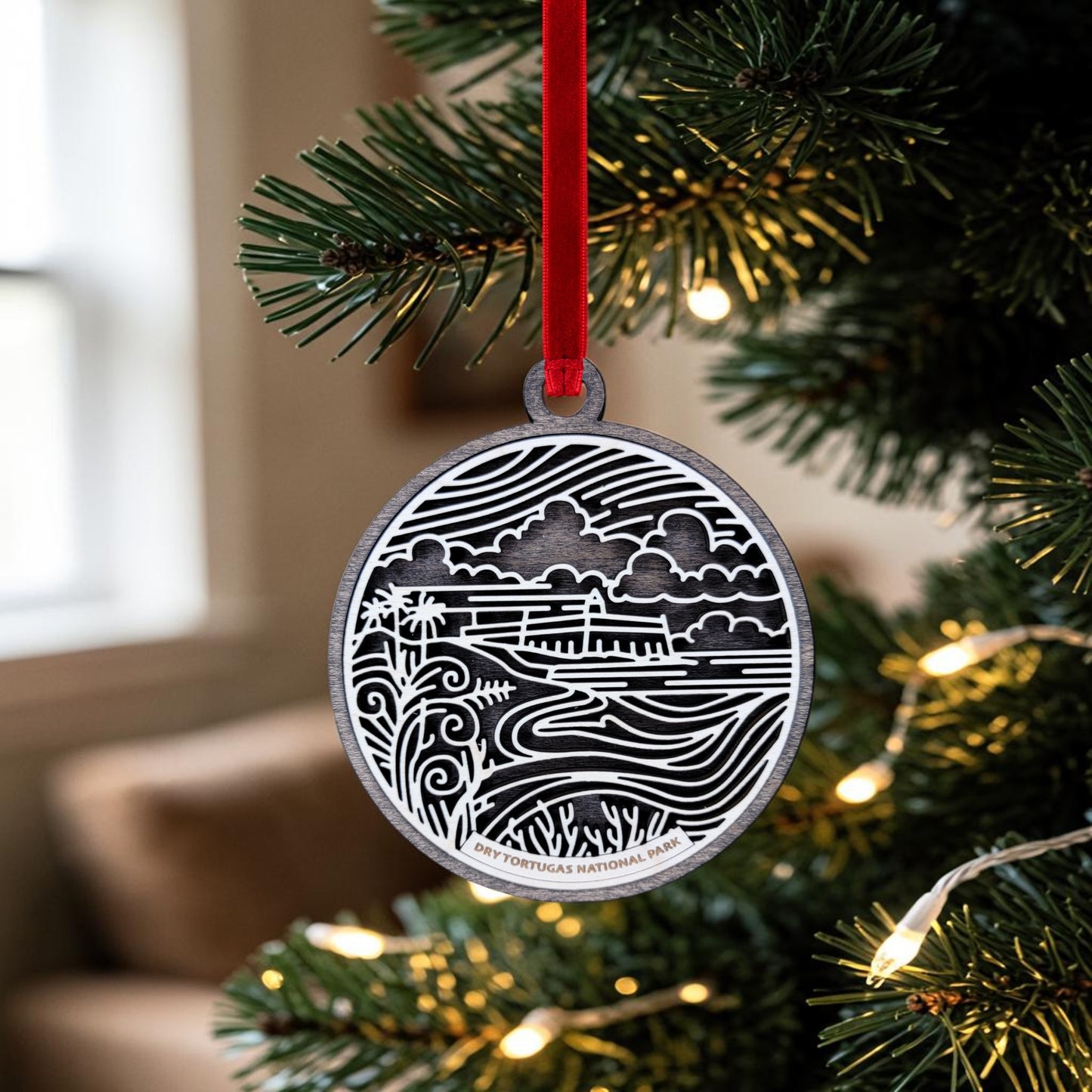 A Mirabilia Boutique Dry Tortugas National Park Christmas Ornament, featuring a black and white coastal scene and the text "DRY TORTUGAS NATIONAL PARK," hangs from a red ribbon on a Christmas tree adorned with lights, with a softly blurred room in the background.