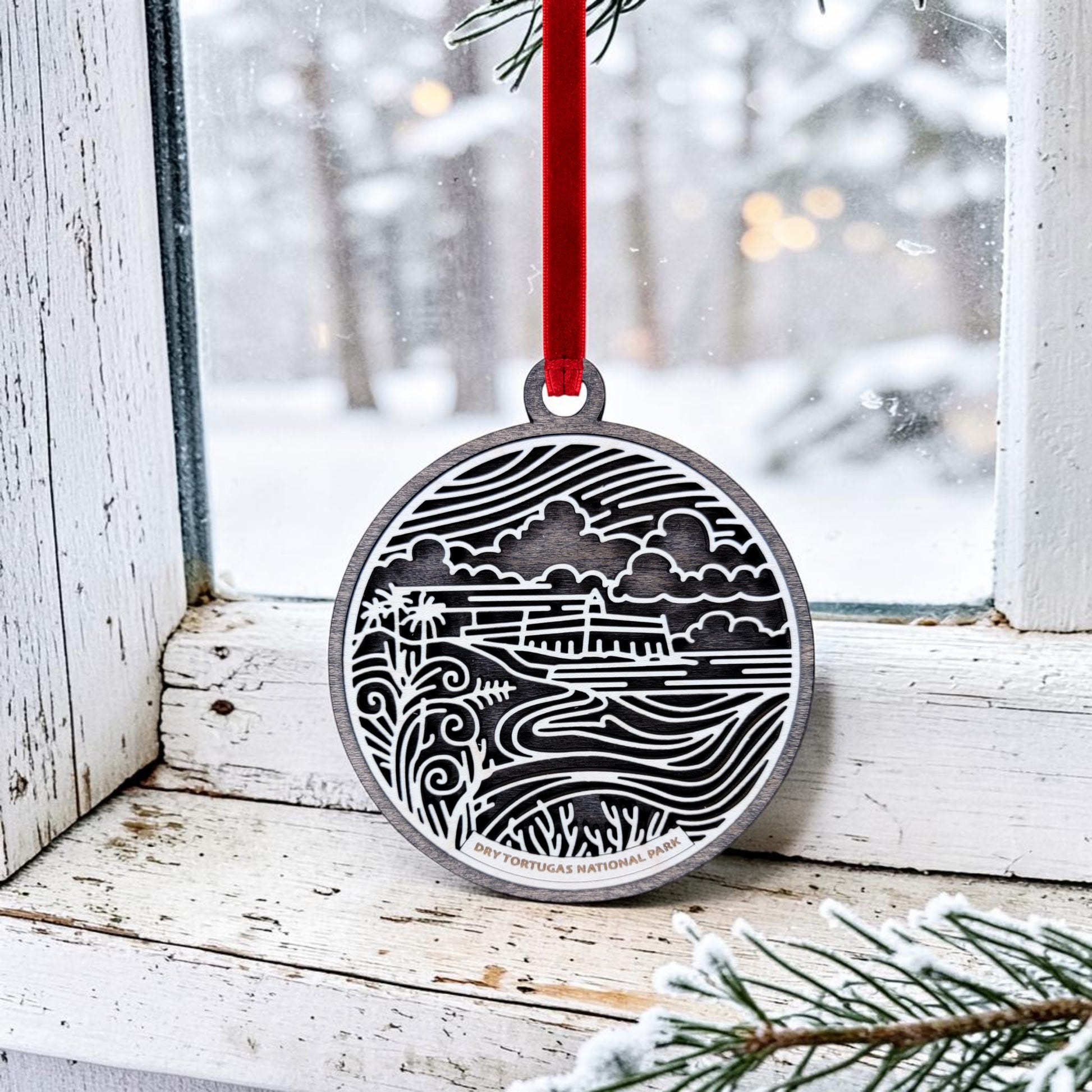 A Mirabilia Boutique Dry Tortugas National Park Christmas Ornament, featuring a silver landscape design with intricate lines illustrating nature and a distant building, hangs on a snowy window by a red ribbon. Frosted pine needles can be seen in the foreground.