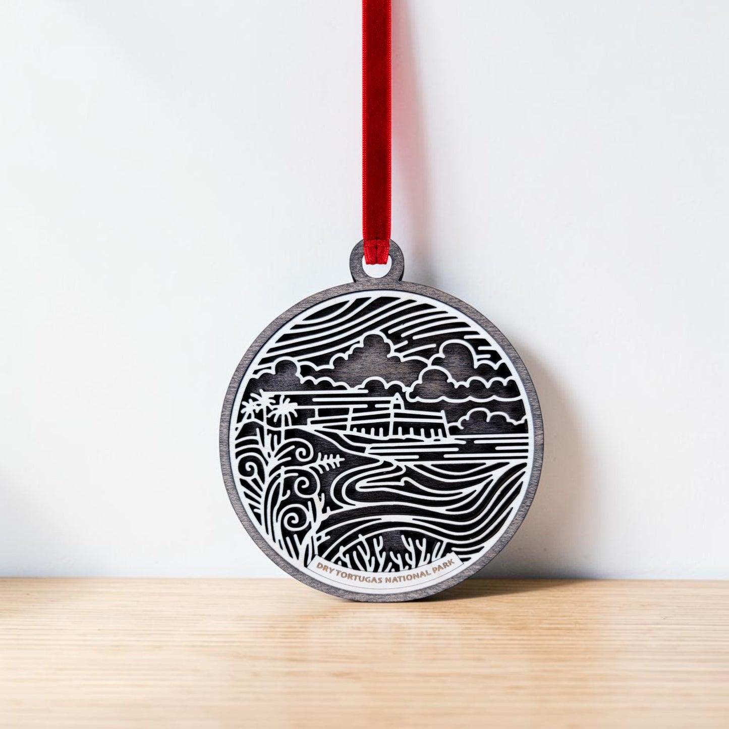 The Mirabilia Boutique Dry Tortugas National Park Christmas Ornament is a round wooden line art ornament engraved with a landscape featuring clouds, a shoreline, plants, and the words "Dry Tortugas National Park." This Fort Jefferson ornament hangs from a red ribbon against a white background and rests on a light wooden surface.