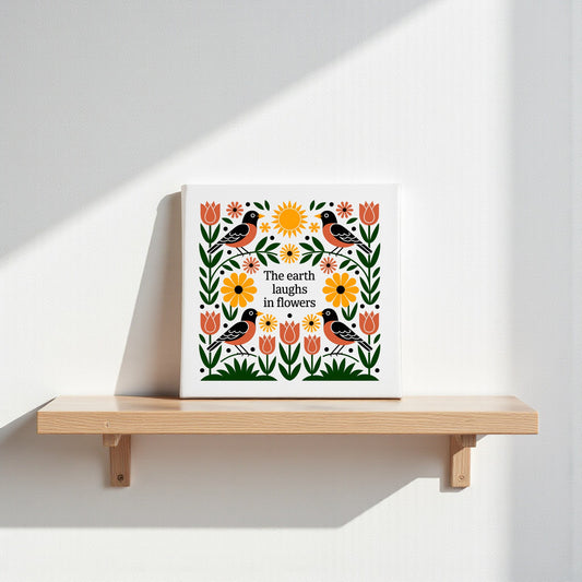 The Earth Laughs in Flowers Canvas Wall Art by Mirabilia Boutique features birds, flowers, and leaves with the quote "The earth laughs in flowers" at the center. Sunlight highlights this charming folk art piece on a wooden shelf.