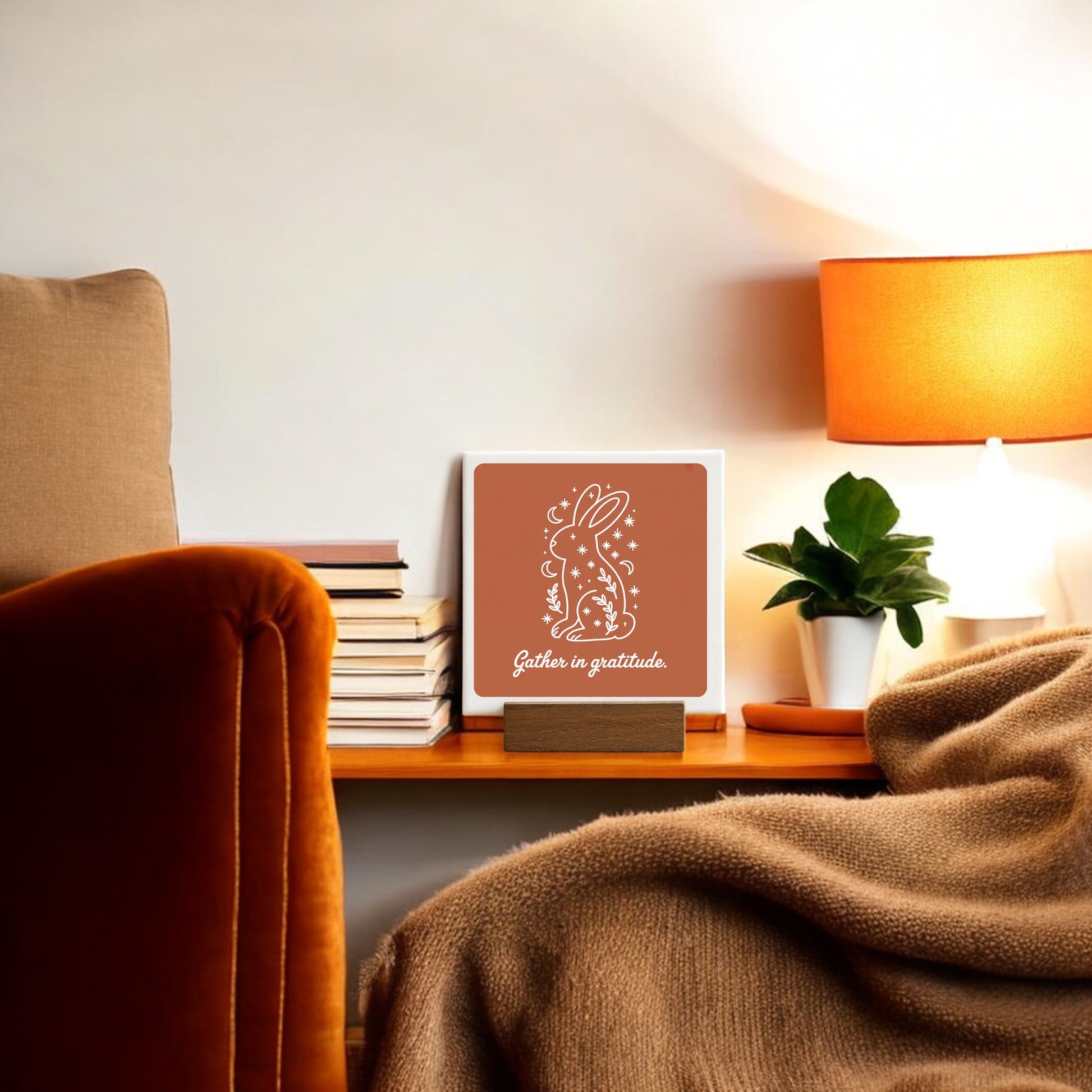 A cozy living space is warmed by a brown blanket on a chair, stacked books, a small plant, an orange lamp, and Mirabilia Boutique’s “Gather in Gratitude Mini Canvas Sign – 4x4 Bunny” as gratitude decor.