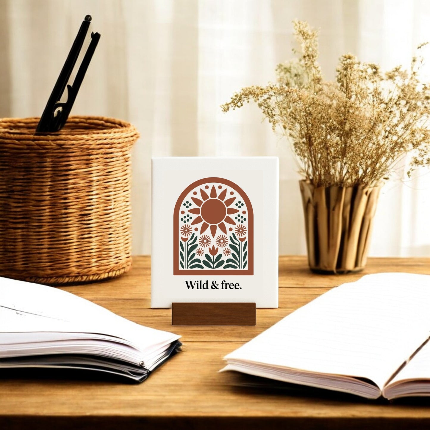 A wooden desk with open notebooks, a pen holder, dried flowers, and the Mirabilia Boutique Wild & Free Mini Sign – Sunflower Boho Desk Art. Sheer curtains let in soft light, enhancing the boho decor and nature-inspired vibe.