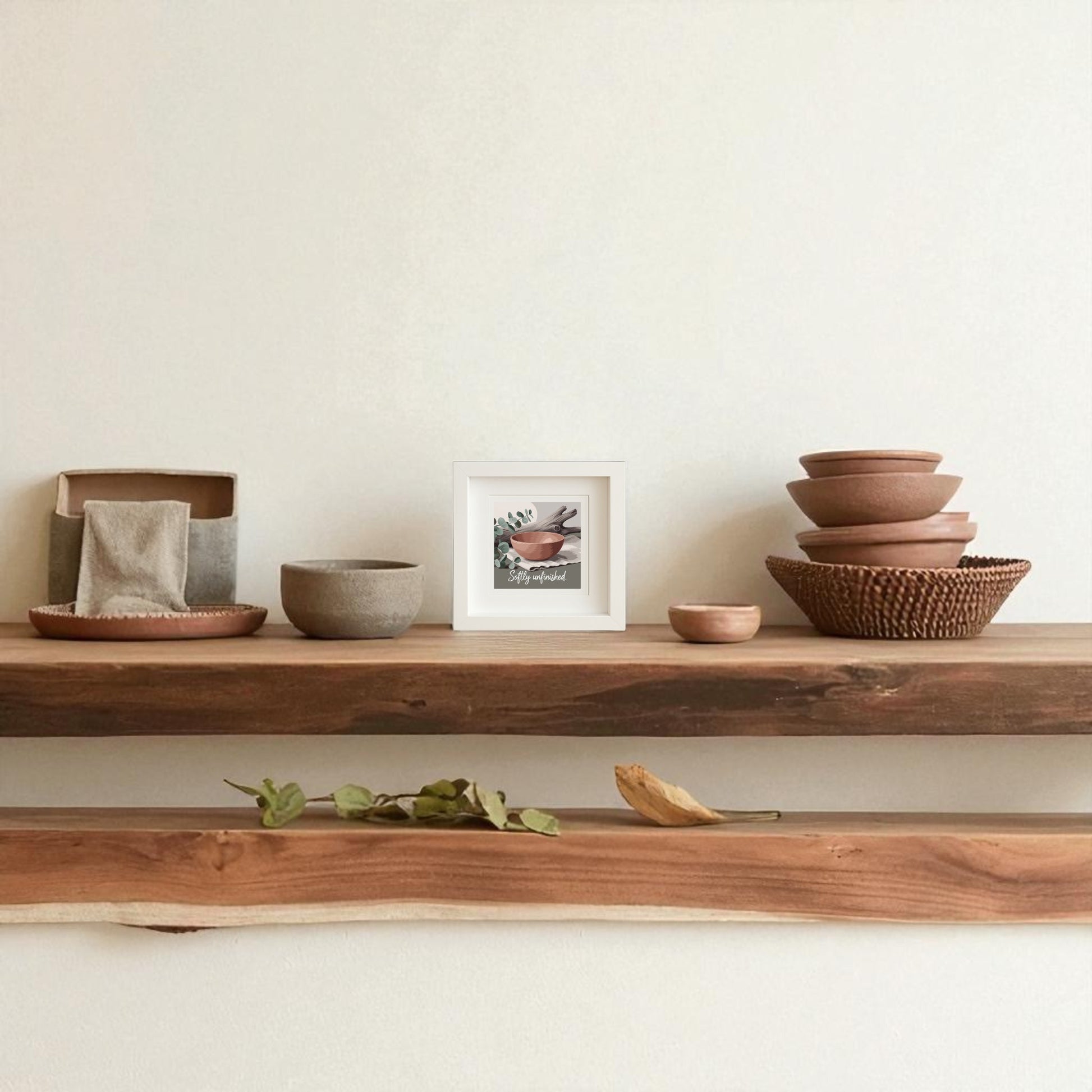 A wooden shelf with stacked ceramic bowls, a small basket, and folded cloth brings modern boho charm. Above, the “Softly Unfinished Shadowbox” Wabi Sabi Boho Wall Art by Mirabilia Boutique is beautifully displayed on a light-colored wall.