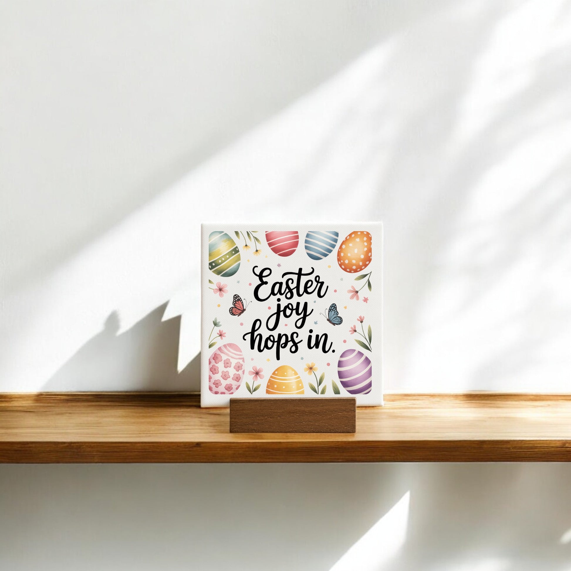 The Mirabilia Boutique Easter Joy Hops In Mini Canvas Sign (4x4) features colorful eggs, flowers, and butterflies with cheerful text, making a charming spring decor piece for your shelf or tabletop.