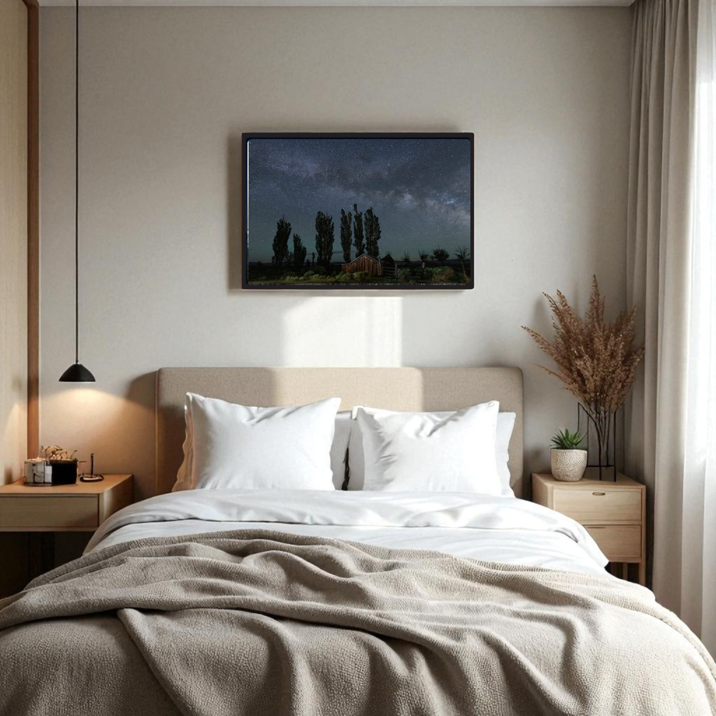 A cozy bedroom with a beige bed, white pillows, two wooden nightstands, a pendant light, and a plant arrangement features the DeChambeau Ranch Milky Way Print by Mirabilia Boutique—an archival-quality framed fine art print of the Eastern Sierra night sky—hung above the bed.