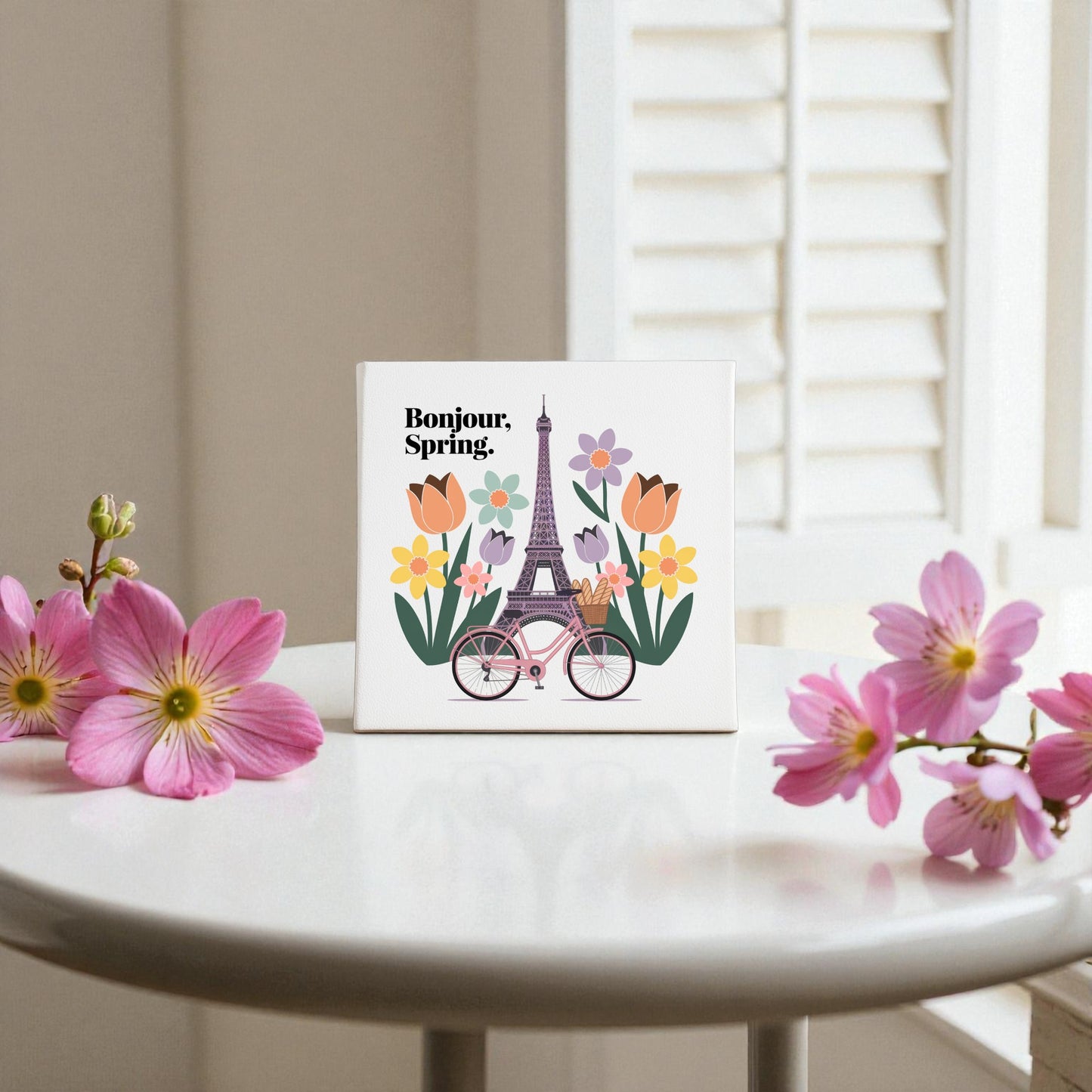 The Bonjour, Spring Canvas – Eiffel Tower Floral Art by Mirabilia Boutique features Eiffel Tower artwork, colorful flowers, a pink bicycle with "Bonjour, Spring" text, and pink flowers in front of white shutters for a charming wall display.