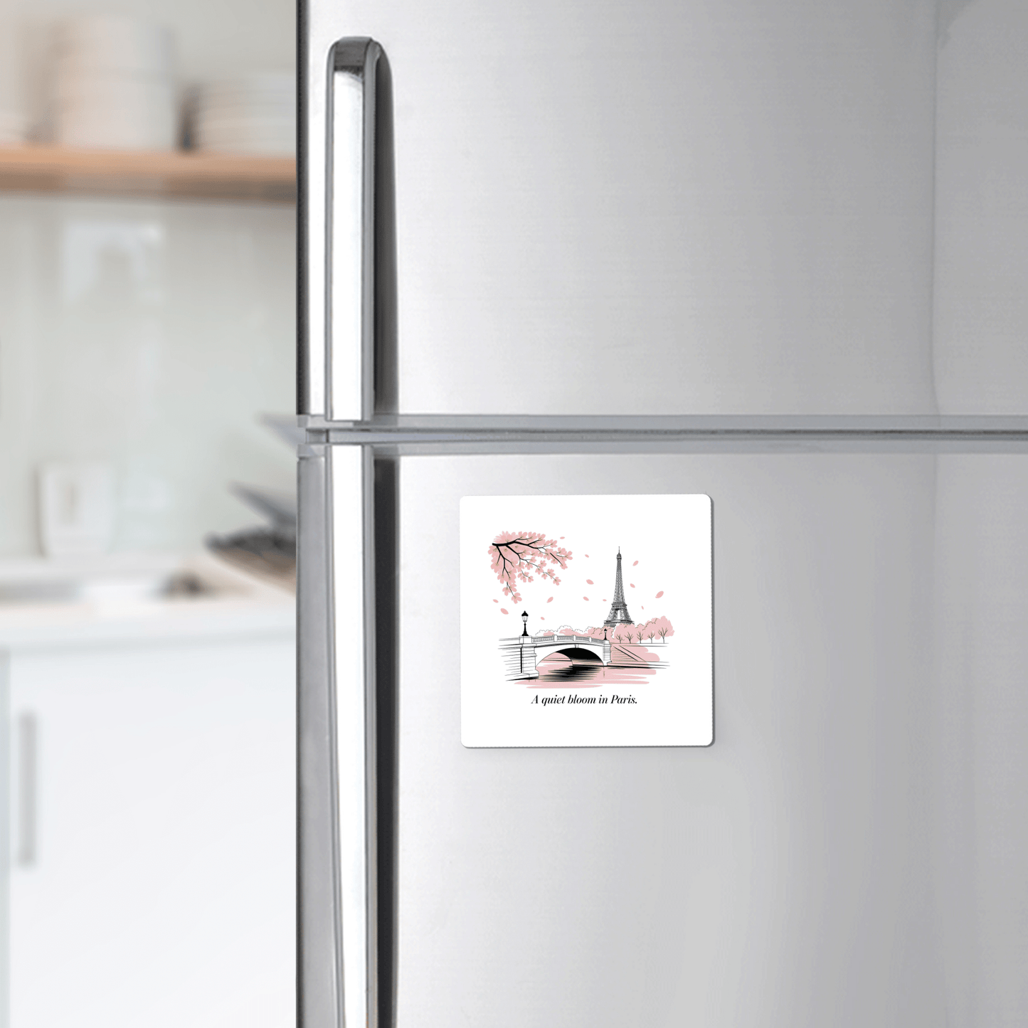 A Quiet Bloom in Paris – Cherry Blossom Magnet by Mirabilia Boutique, featuring the Eiffel Tower, a bridge, pink leaves, and the phrase "A quiet blossom in Paris," is shown on a refrigerator with a kitchen counter and shelves in the background.