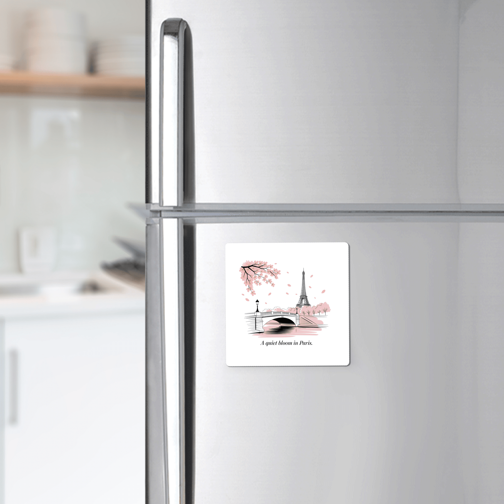 A Quiet Bloom in Paris – Cherry Blossom Magnet by Mirabilia Boutique, featuring the Eiffel Tower, a bridge, pink leaves, and the phrase "A quiet blossom in Paris," is shown on a refrigerator with a kitchen counter and shelves in the background.