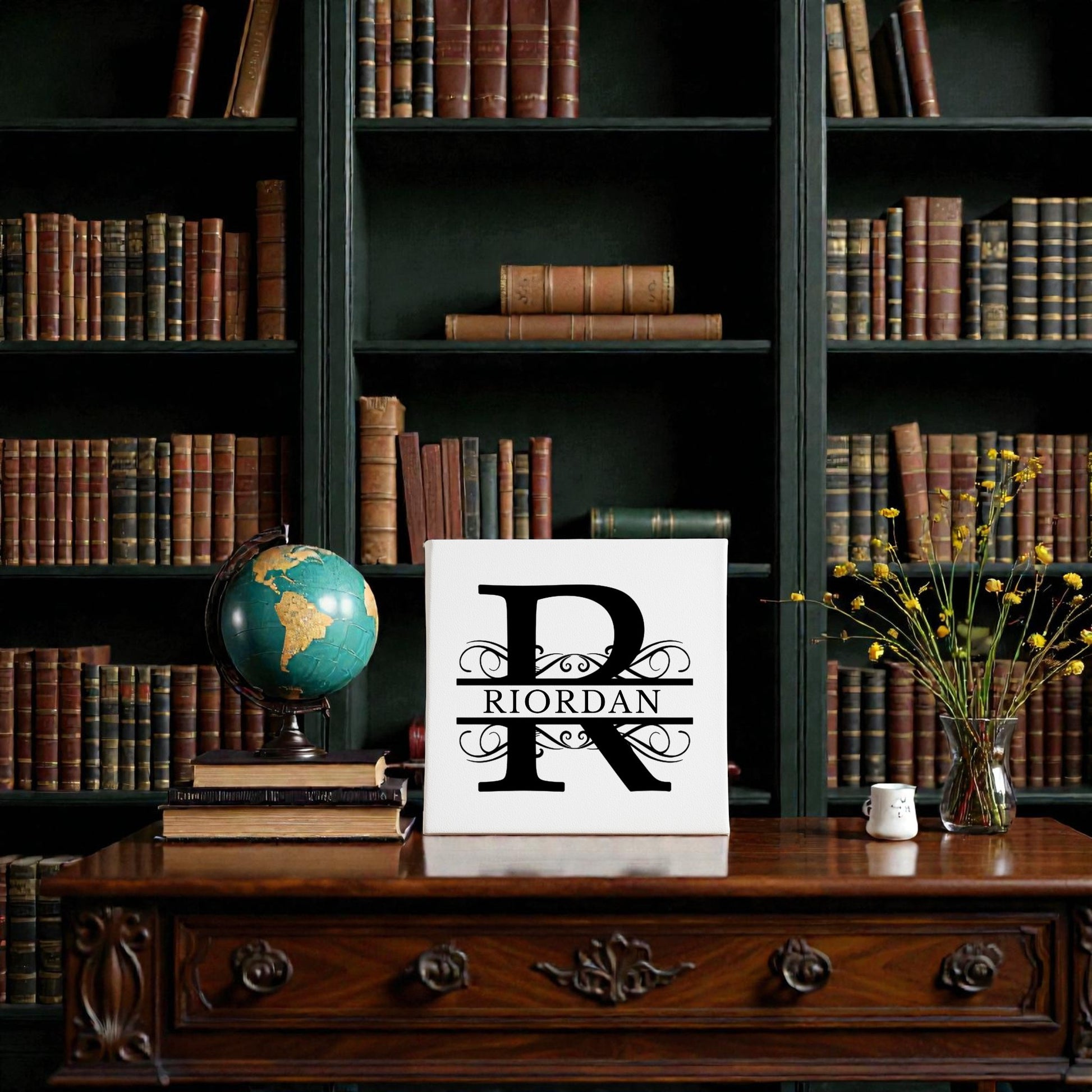 A wooden desk sits before a dark bookcase lined with old books. On the desk are a vintage globe, yellow flowers, and a Mirabilia Boutique Custom Monogram Canvas with Family Name – 8x8, personalized for Riordan.