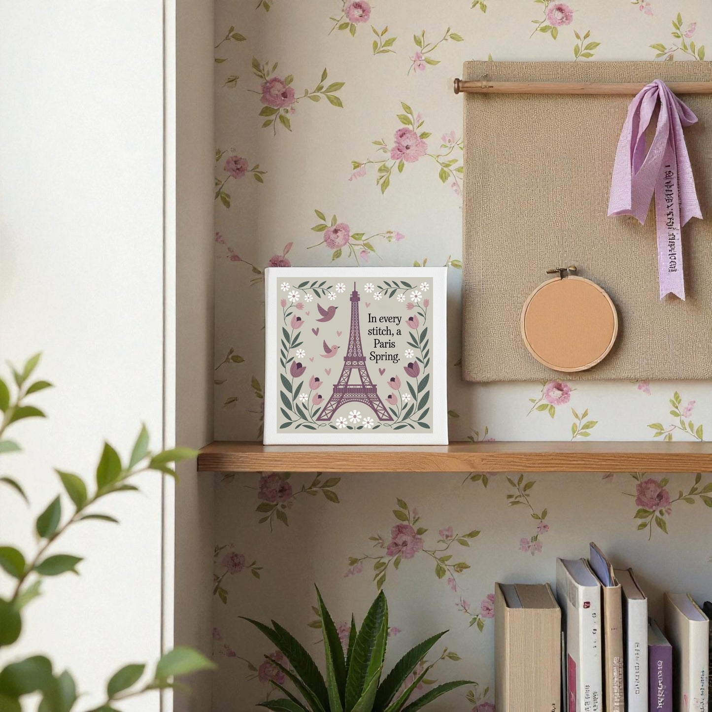 A shelf displays books, a potted plant, and the Mirabilia Boutique Paris Spring Stitch Canvas—embroidered-style floral art with birds and “In every sketch, a Paris Spring”—set against floral wallpaper and cross-stitch-inspired decor.