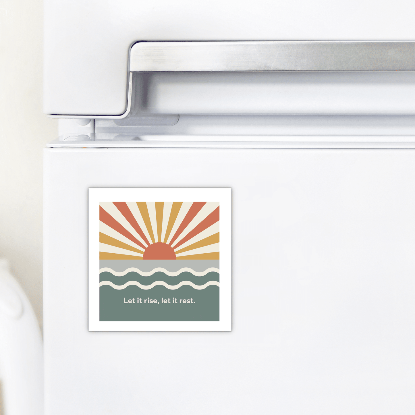 The Let It Rise, Let It Rest Magnet – Sunrise Affirmation by Mirabilia Boutique features a stylized sun and wavy water with warm hues, adding an uplifting, inspirational accent to your kitchen.