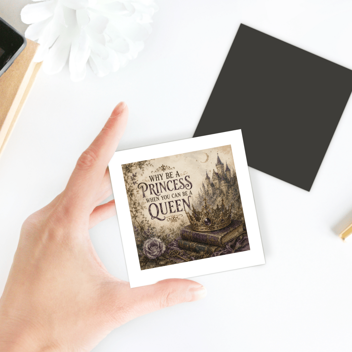 A hand holds the "Why Be a Princess When You Can Be a Queen" magnet by Mirabilia Boutique, featuring an illustration of a castle and books. Ideal for fairytale decor lovers, other stationery items are visible on a white desk in the background.