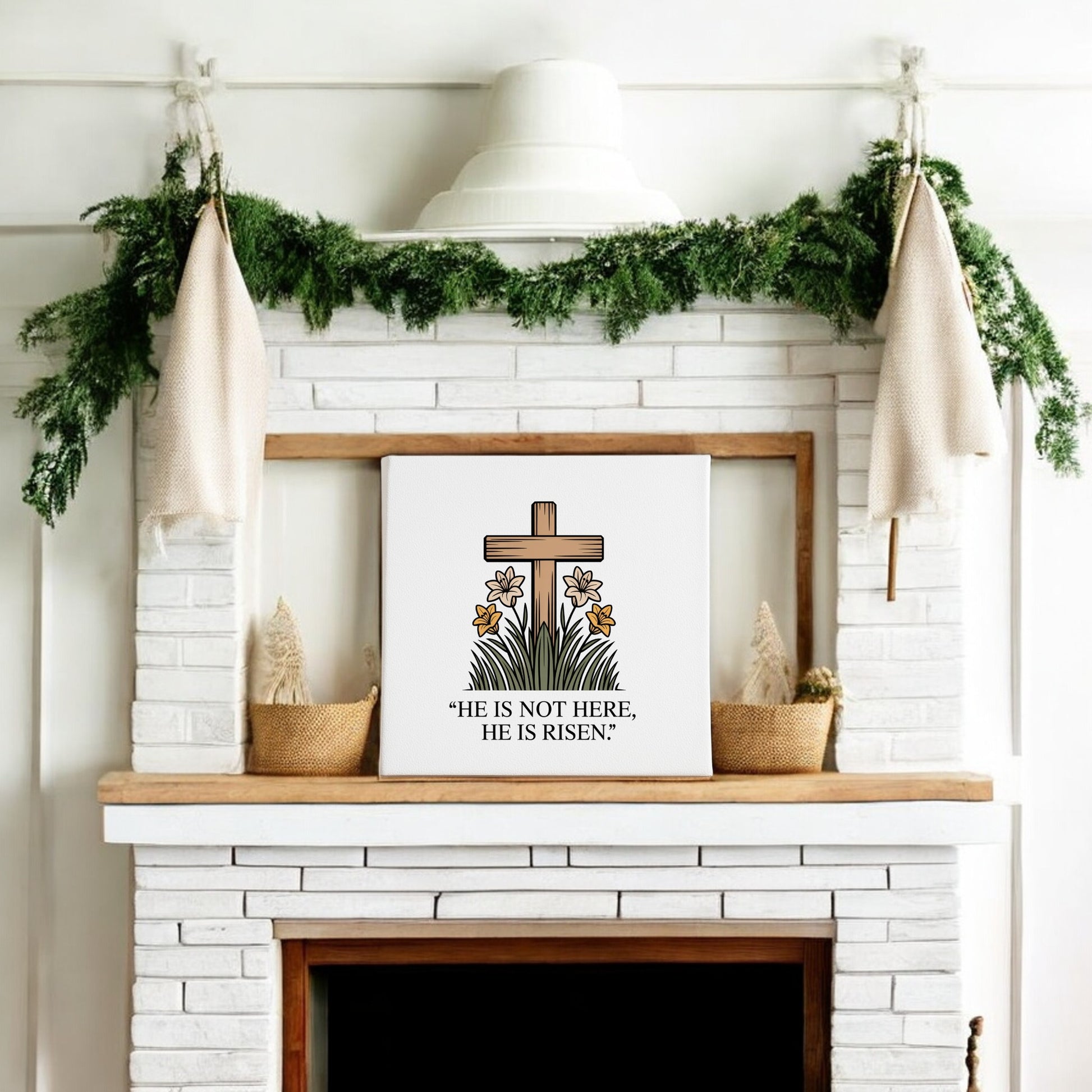 A white brick fireplace decorated with greenery, neutral stockings, and an 8x8 “He Is Risen Cross & Lily Canvas” by Mirabilia Boutique—featuring a cross, lilies, and the Easter message “He is not here, He is risen.”.