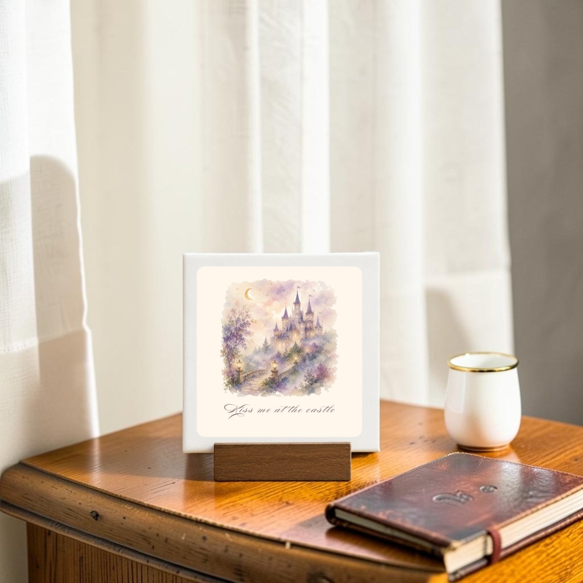 The Kiss Me at the Castle Mini Canvas Sign by Mirabilia Boutique features a whimsical castle scene and the words “Kiss Me at the Castle.” Displayed on a wooden stand beside a cup and a closed notebook, it adds a charming fairytale touch to your table near a sunlit window with white curtains.