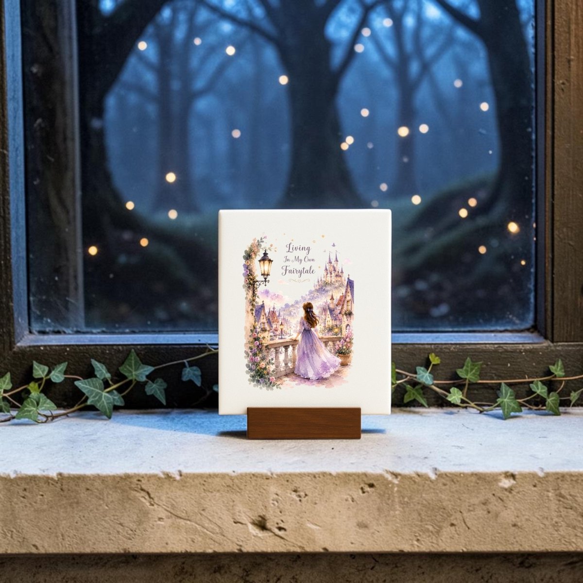 The Living in My Own Fairytale Mini Canvas by Mirabilia Boutique features an illustration of a woman in a purple dress near a castle, with the words "Living My Own Fairytale." Displayed on a windowsill adorned with ivy and set against a window revealing a dark, tree-filled night lit by glowing lights, this charming piece is perfect whimsical wall art for anyone who loves fairytale decor.