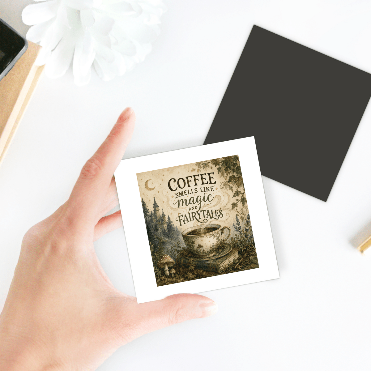 A hand holds the Mirabilia Boutique “Coffee Smells Like Magic And Fairytales” magnet, featuring an illustrated coffee cup and whimsical lettering. Perfect as a charming coffee gift or a magical accent for your fairy tale kitchen decor, the magnet is photographed on a white table with a black square and flower detail in the background.