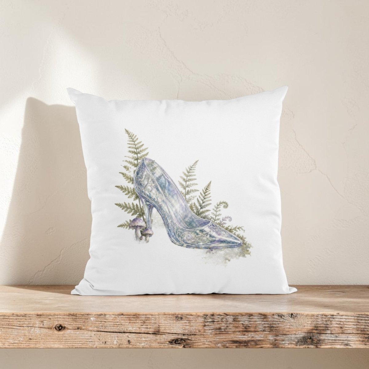 Add fairytale charm to your home décor with the Enchanted Glass Slipper Throw Pillow by Mirabilia Boutique—a white woodland pillow featuring a watercolor-style high-heeled shoe surrounded by delicate ferns and a small mushroom, elegantly displayed on a wooden shelf against a light textured wall.