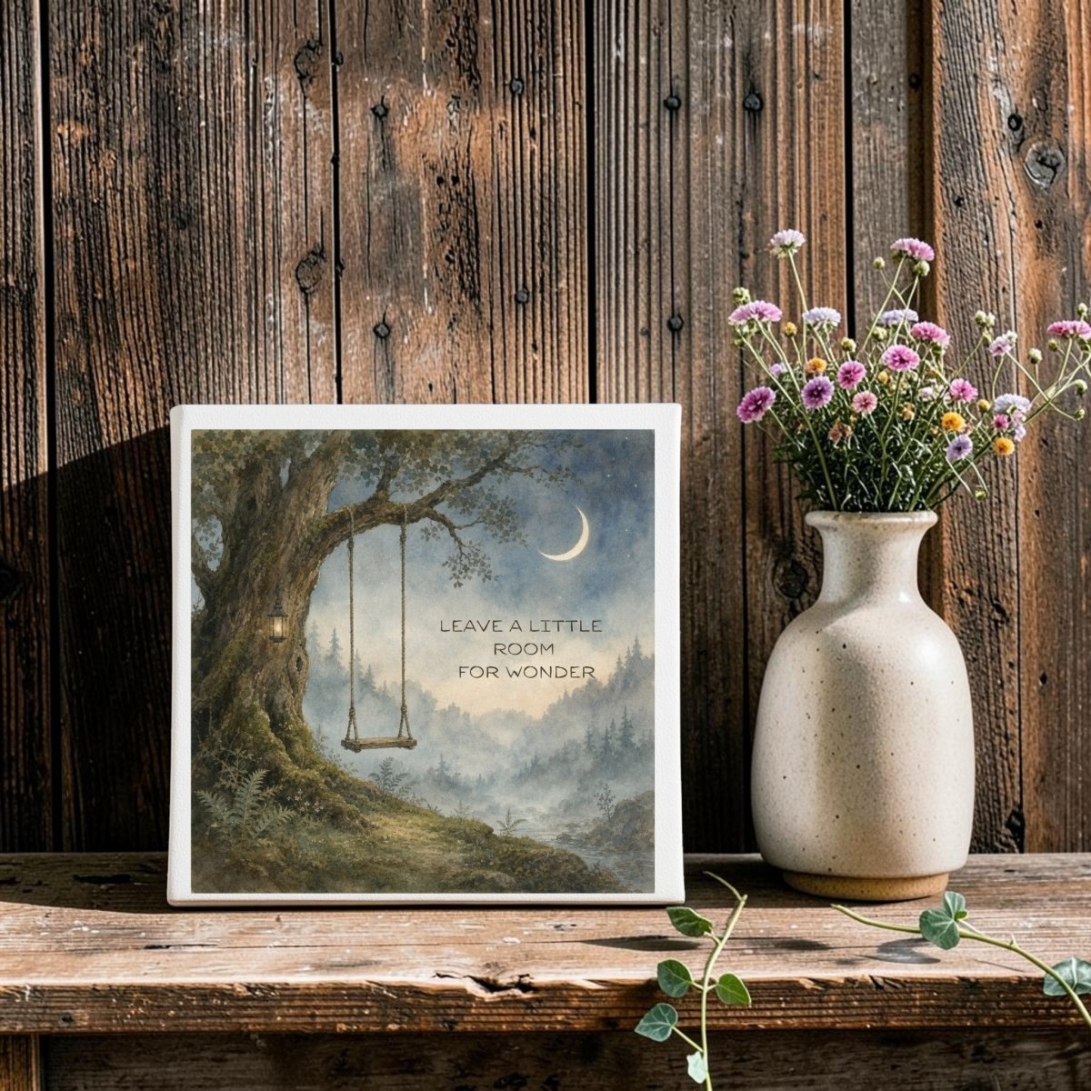 The Leave a Little Room for Wonder Canvas by Mirabilia Boutique features a watercolor landscape of a tree swing beneath a crescent moon, with the phrase "Leave a little room for wonder." Displayed on a wooden shelf beside a beige vase holding purple and white flowers, this nature-inspired art piece brings rustic charm against its wooden wall backdrop.