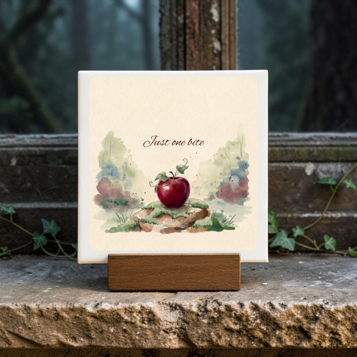 The Just One Bite Mini Canvas Sign by Mirabilia Boutique features a watercolor illustration of a red apple and the words "Just one bite." Displayed on a wooden stand in front of an old window with plants around the stone sill, it makes for charming fairy tale decor.