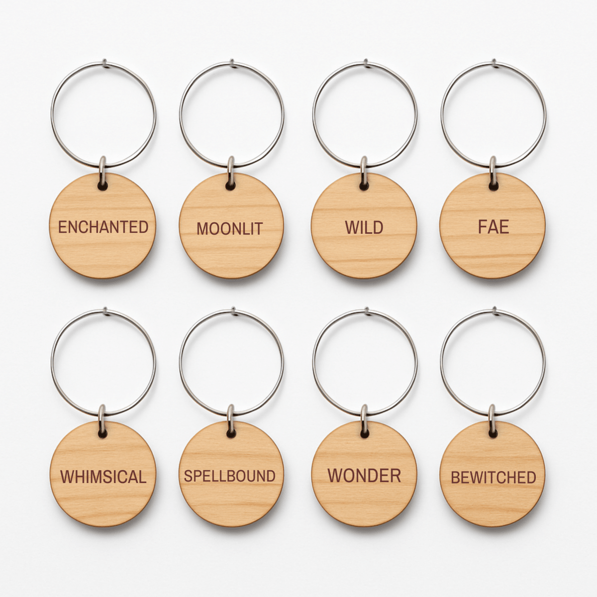 The Enchanted Woodland Wine Charms Set by Mirabilia Boutique features eight round wooden keychains with metal rings, each engraved with a unique word—ENCHANTED, MOONLIT, WILD, FAE, WHIMSICAL, SPELLBOUND, WONDER, and BEWITCHED. Arranged in two neat rows on a white background, these woodland wine charms are perfect as enchanting wine glass markers and bring a whimsical touch to any gathering.