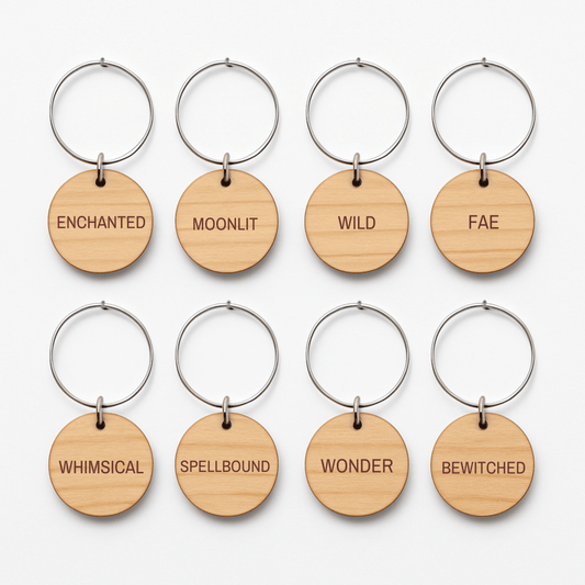 The Enchanted Woodland Wine Charms Set by Mirabilia Boutique features eight round wooden keychains with metal rings, each engraved with a unique word—ENCHANTED, MOONLIT, WILD, FAE, WHIMSICAL, SPELLBOUND, WONDER, and BEWITCHED. Arranged in two neat rows on a white background, these woodland wine charms are perfect as enchanting wine glass markers and bring a whimsical touch to any gathering.
