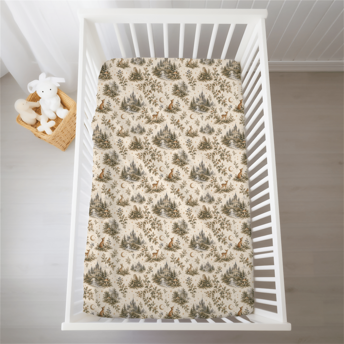 A white crib with a mattress dressed in the Enchanted Woodlands Crib Sheet by Mirabilia Boutique, featuring mesmerizing trees, foxes, and moons inspired by enchanted forests. Beside the crib on a light wood floor sits a plush bunny nestled in a wicker basket.