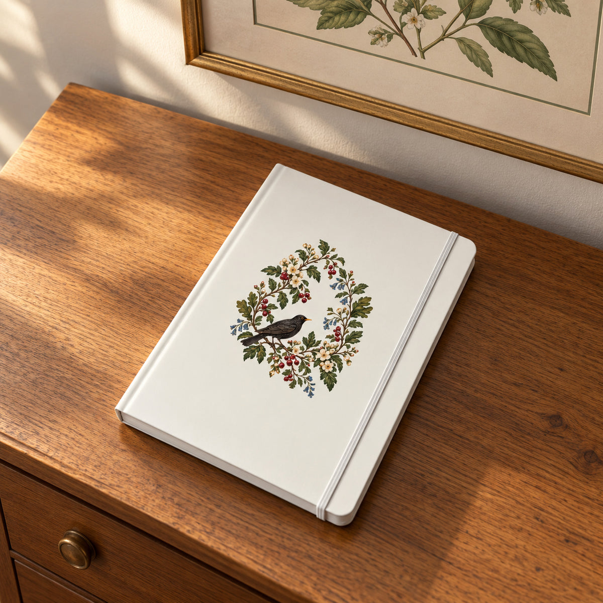 The Garden Ledger A5 Lined Journal by Mirabilia Boutique, a cream-colored journal featuring a blackbird perched within a botanical wreath of flowers and berries, resting on a wooden surface.
