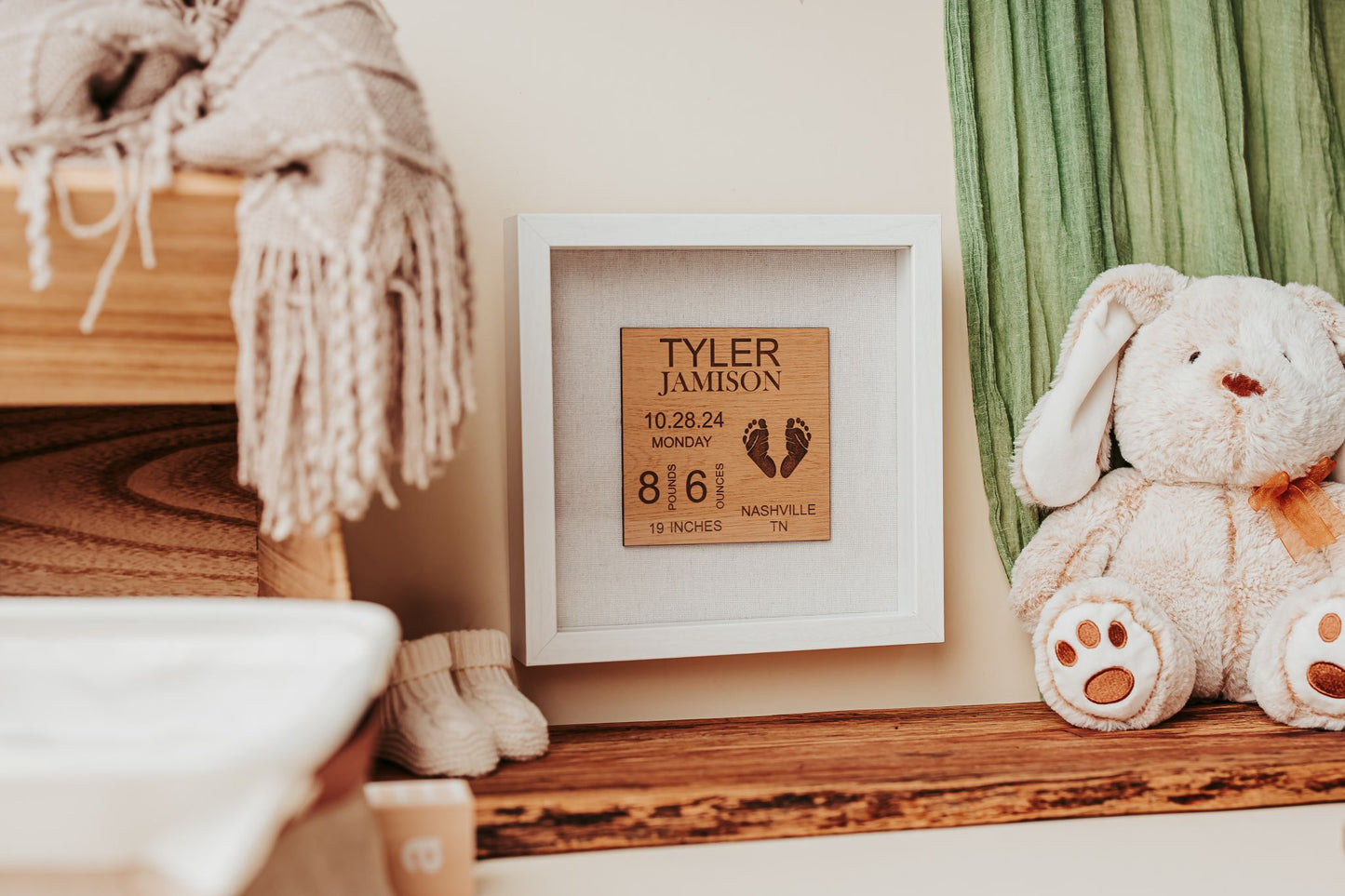 The Mirabilia Boutique Personalized Baby Birth Stats Shadowbox, a 9x9 laser engraved nursery keepsake with custom name and stats, sits on a wooden shelf beside a stuffed bunny, folded blanket, and green curtain in a cozy nursery.