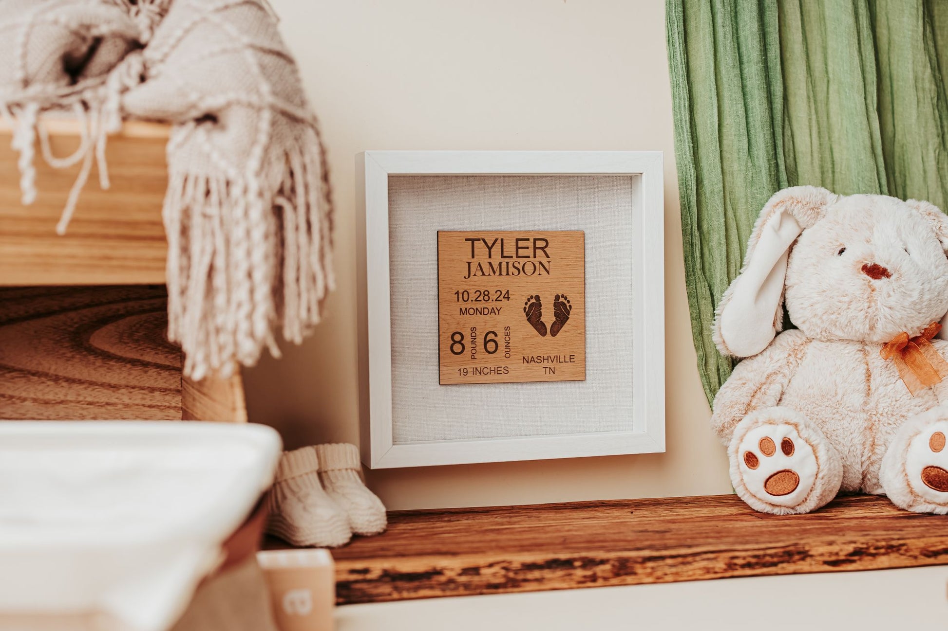 The Mirabilia Boutique Personalized Baby Birth Stats Shadowbox, a 9x9 laser engraved nursery keepsake with custom name and stats, sits on a wooden shelf beside a stuffed bunny, folded blanket, and green curtain in a cozy nursery.
