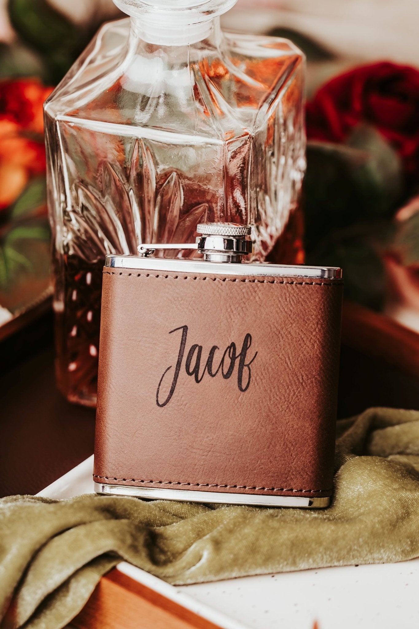 The Custom Engraved Flask by Mirabilia Boutique, a personalized leather gift for him, sits in front of a crystal decanter with amber liquid, surrounded by flowers and green cloth—perfect as a groomsmen gift.