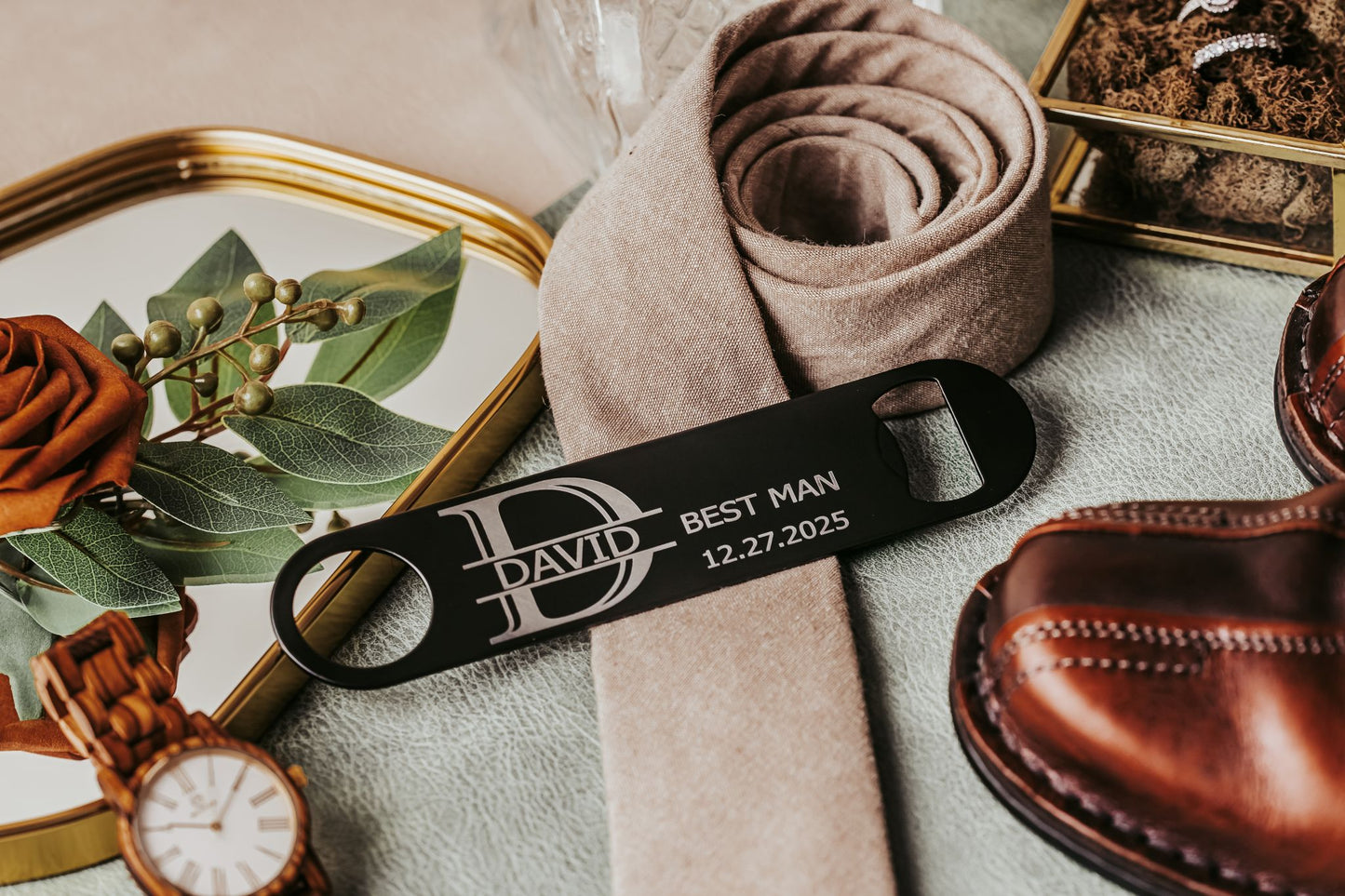 A flatlay featuring a watch, dress shoes, beige tie, decorative tray with a rose, and the Mirabilia Boutique Engraved Flat Metal Bottle Opener personalized for “David, Best Man, 12.27.2025,” displayed on a textured surface.