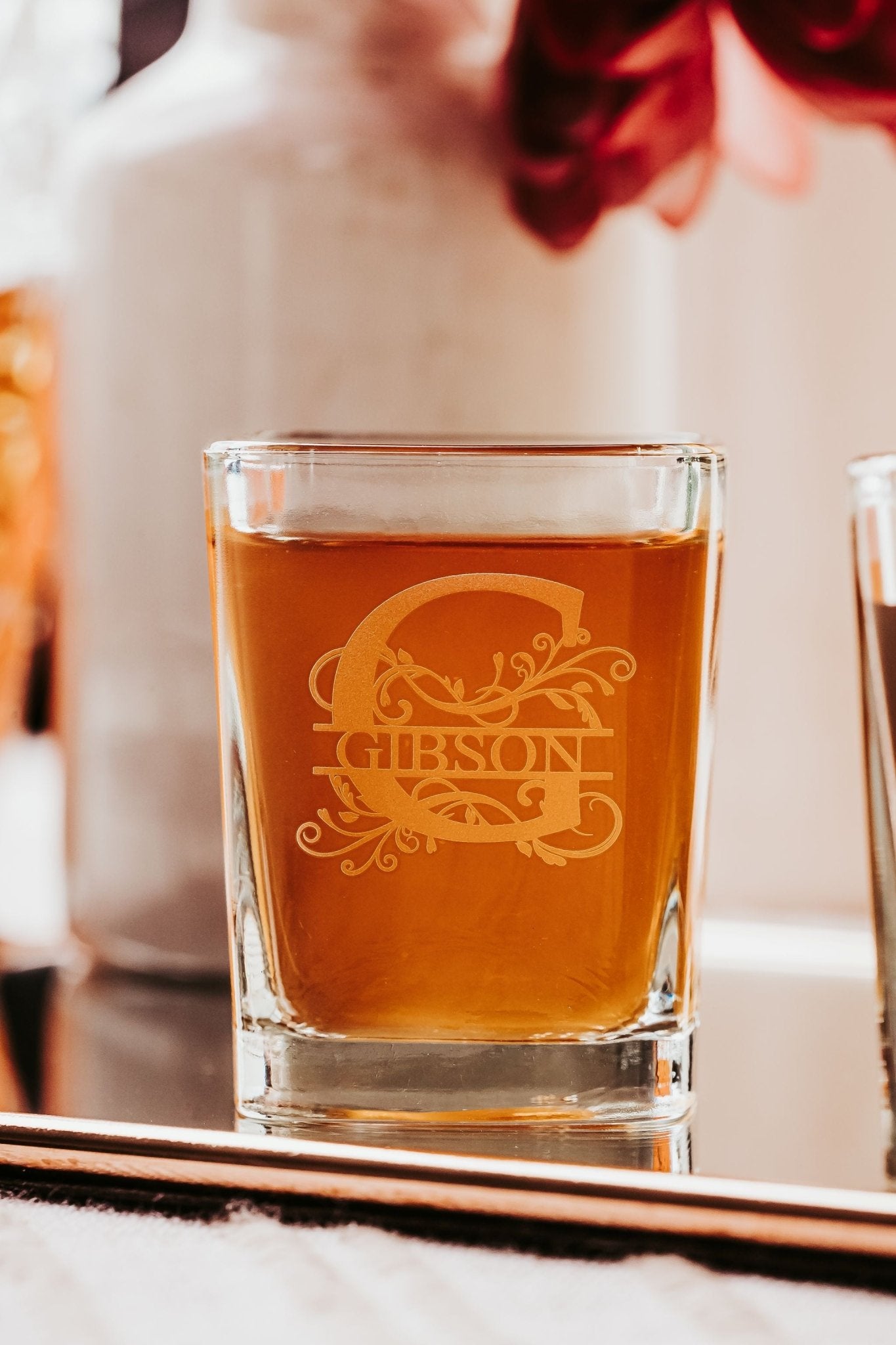 A Personalized Monogram Shot Glass from Mirabilia Boutique, etched with an ornate "G" and "Gibson," sits on a reflective surface, filled with amber liquid. This custom engraved gift set makes a unique, elegant addition to any collection.