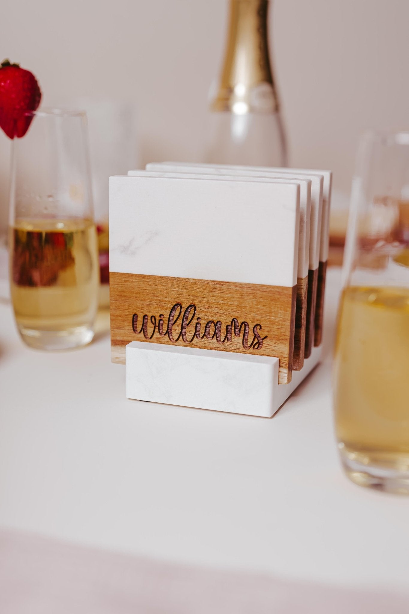 A set from Mirabilia Boutique's Custom Engraved Marble and Acacia Coaster collection, featuring elegantly engraved coasters and a personalized wooden holder with the name "Williams," sits on a white table surrounded by glasses filled with a light golden beverage.
