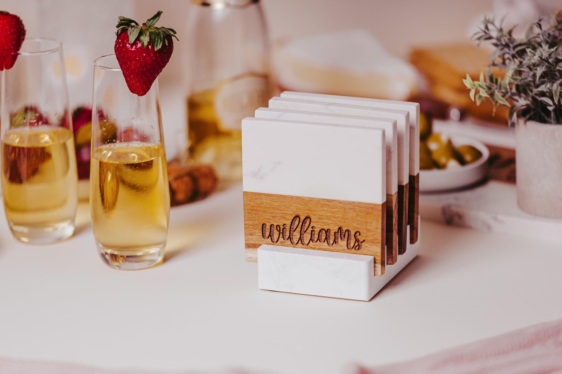 A set of Custom Engraved Marble and Acacia Coasters from Mirabilia Boutique, personalized with the name "Williams," is elegantly displayed on their matching coaster stand. Adjacent are two glasses filled with sparkling beverages accented by strawberries, along with a bottle, a dish of olives, and a decorative plant set on a white surface.