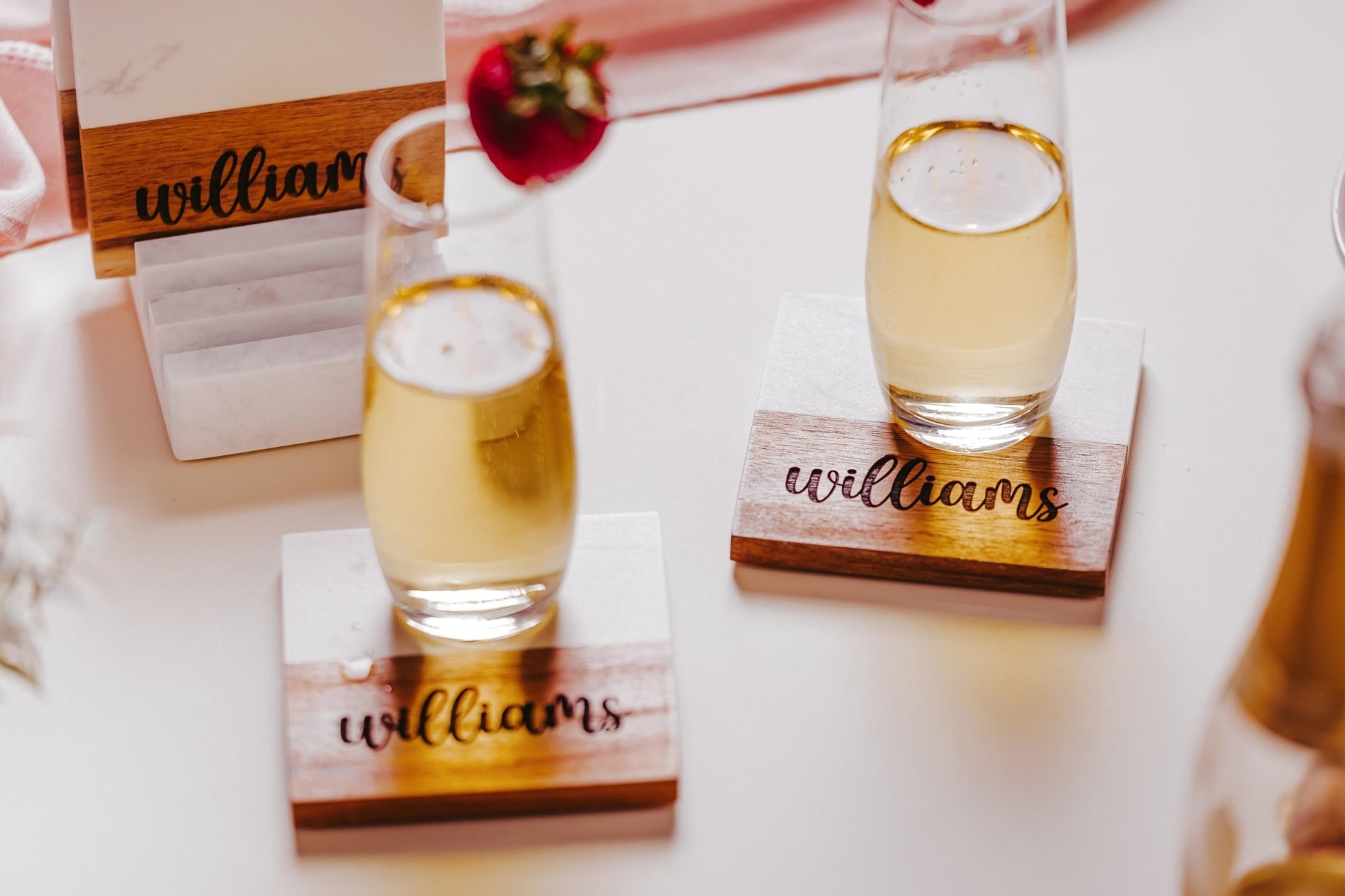Two glasses of champagne sit elegantly on the Mirabilia Boutique Custom Engraved Marble and Acacia Coaster Set, featuring the name "Williams." One of the glasses is adorned with a strawberry on its rim, while in the background, an exquisite wooden holder showcases name cards, enhancing the festive atmosphere.