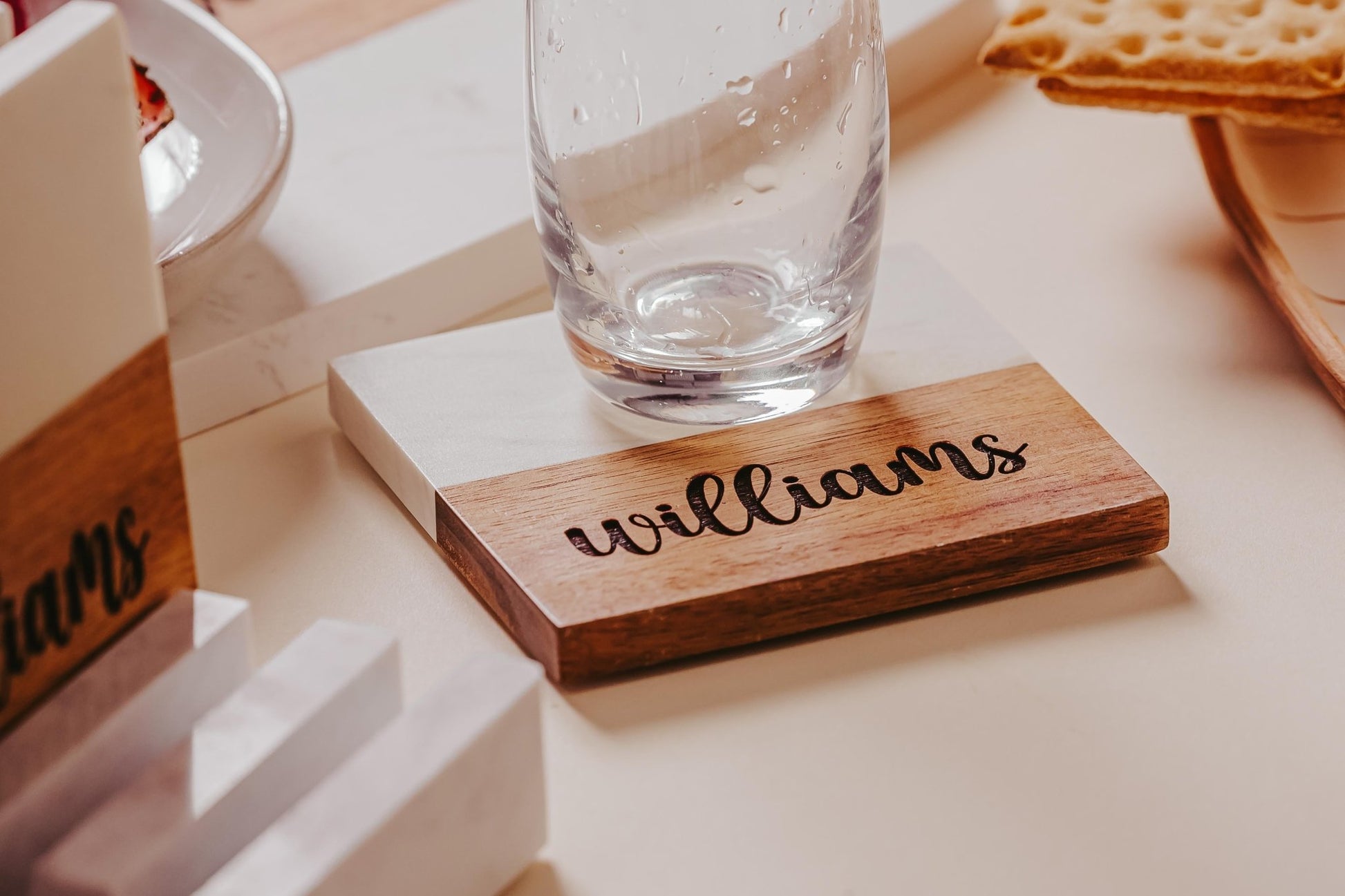 A personalized coaster featuring "Williams" in cursive script supports a clear, empty glass, creating an elegant display on a light-colored table. This setup includes crackers and decorative items, all enhanced by the Custom Engraved Marble and Acacia Coaster Set from Mirabilia Boutique for added sophistication.