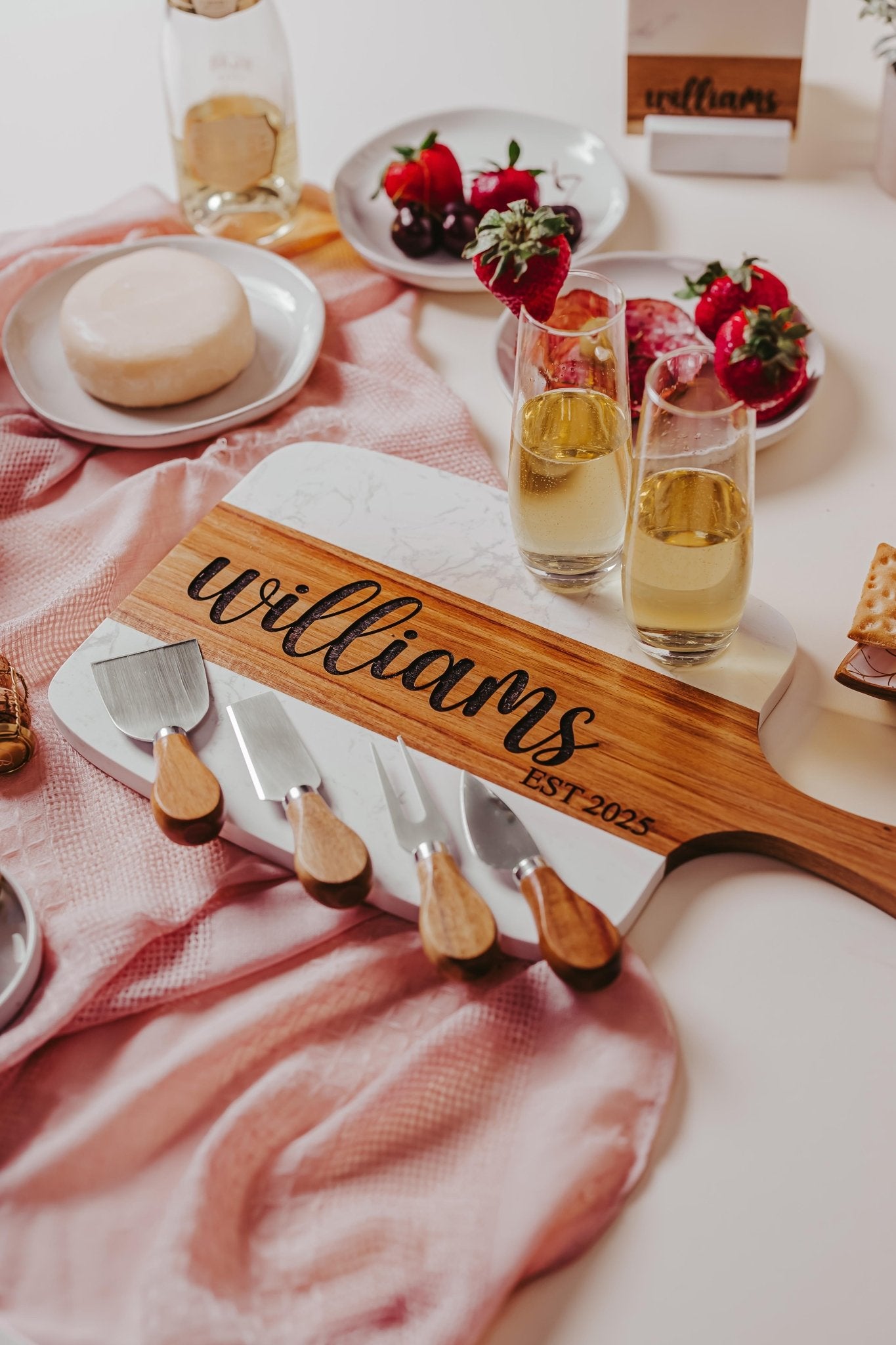 A unique keepsake, the Personalized Engraved Marble and Acacia Serving Board by Mirabilia Boutique features the engraving "williams EST. 2025" and comes with three cheese knives. Nearby, champagne glasses, strawberries, cheese, and crackers are artfully arranged on a pink cloth.