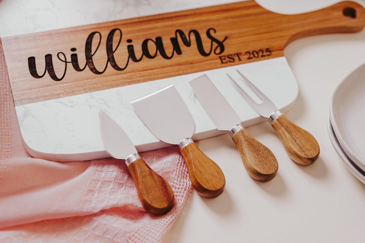 Mirabilia Boutique's Personalized Engraved Marble and Acacia Serving Board features the engraving "Williams EST 2025" and is complete with four cheese knives featuring wooden handles. A pink cloth partially drapes over this custom keepsake, with a silver ribbon peeking out on the right.