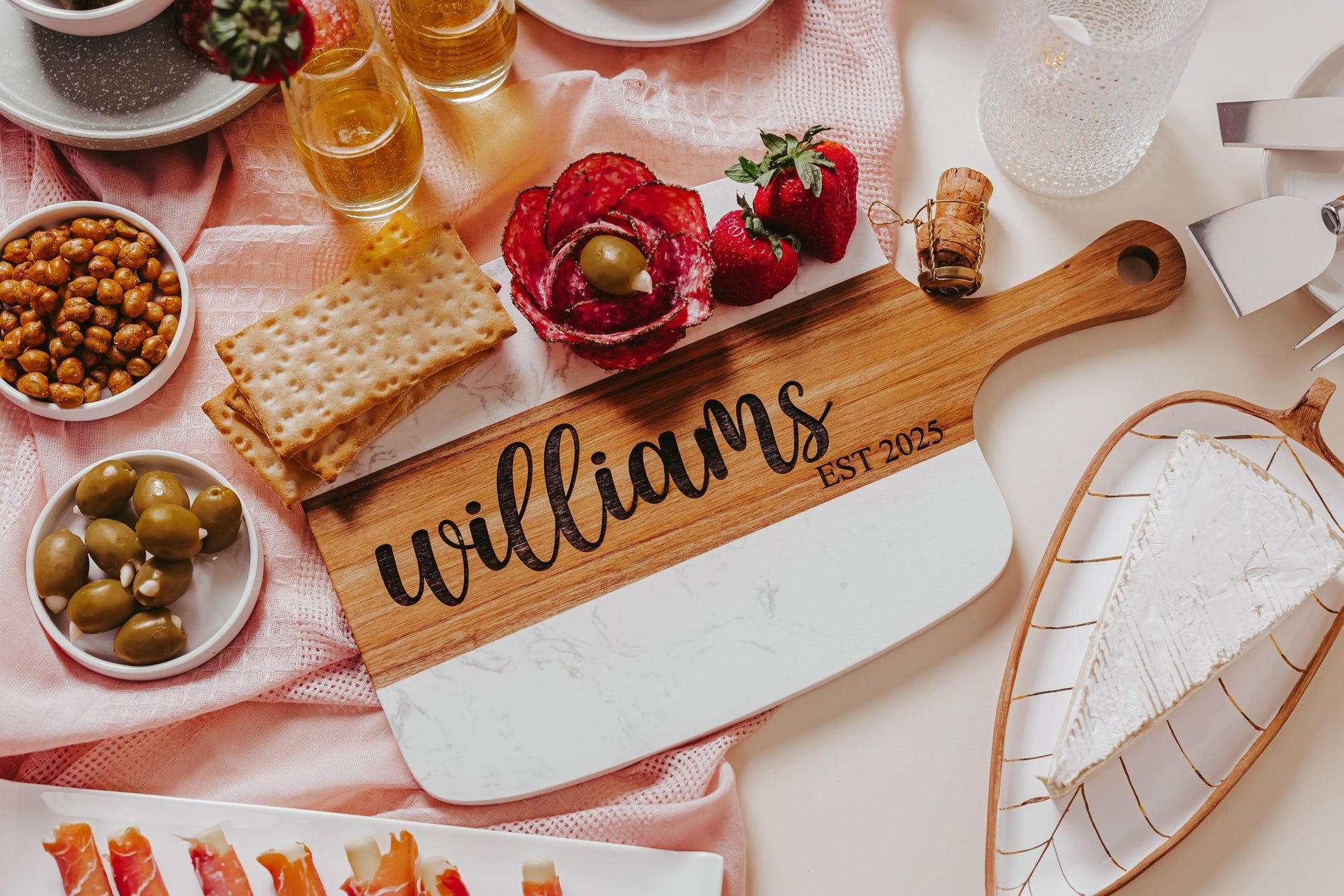 A Personalized Engraved Marble and Acacia Serving Board from Mirabilia Boutique, featuring the engravings "Williams" and "Est 2025," is elegantly arranged with crackers, meats, olives, strawberries, cocktail glasses, a cheese wedge, and snacks on a pink cloth.