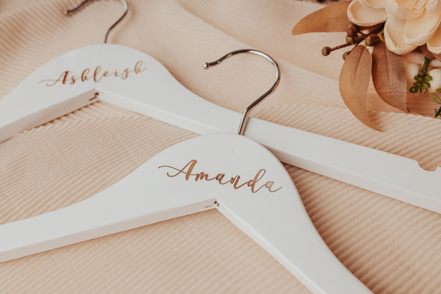 The Personalized Wedding Hangers | Custom Name Hanger Set by Mirabilia Boutique features custom names in elegant gold script on wood, displayed on beige fabric with cream and brown faux flowers—ideal for capturing your special wedding day details.