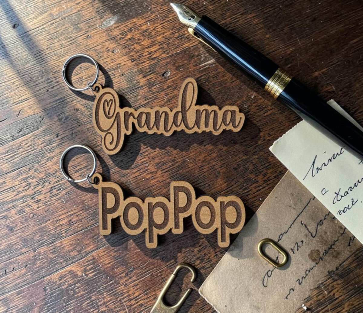 Two Personalized Engraved Wood Name Keychains by Mirabilia Boutique, customized with "Grandma" and "PopPop," rest on a wooden table beside a black fountain pen, an old letter, and a gold paperclip, while sunlight casts soft shadows across the scene.