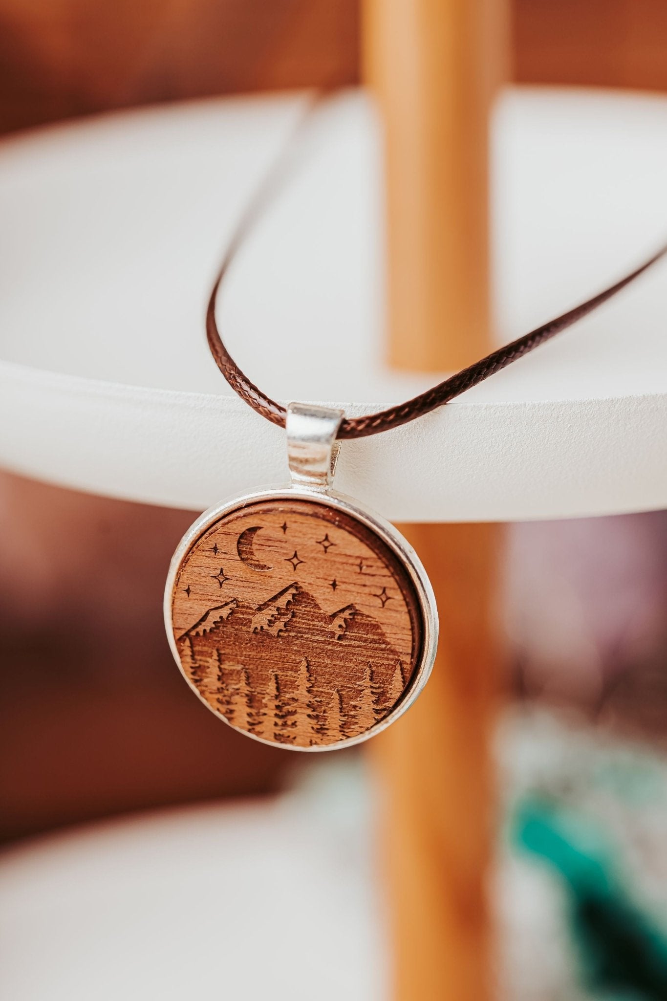 The Mirabilia Boutique Engraved Wood Pendant Necklace features a round mountain night sky design and is displayed on a white stand—perfect for boho jewelry lovers.